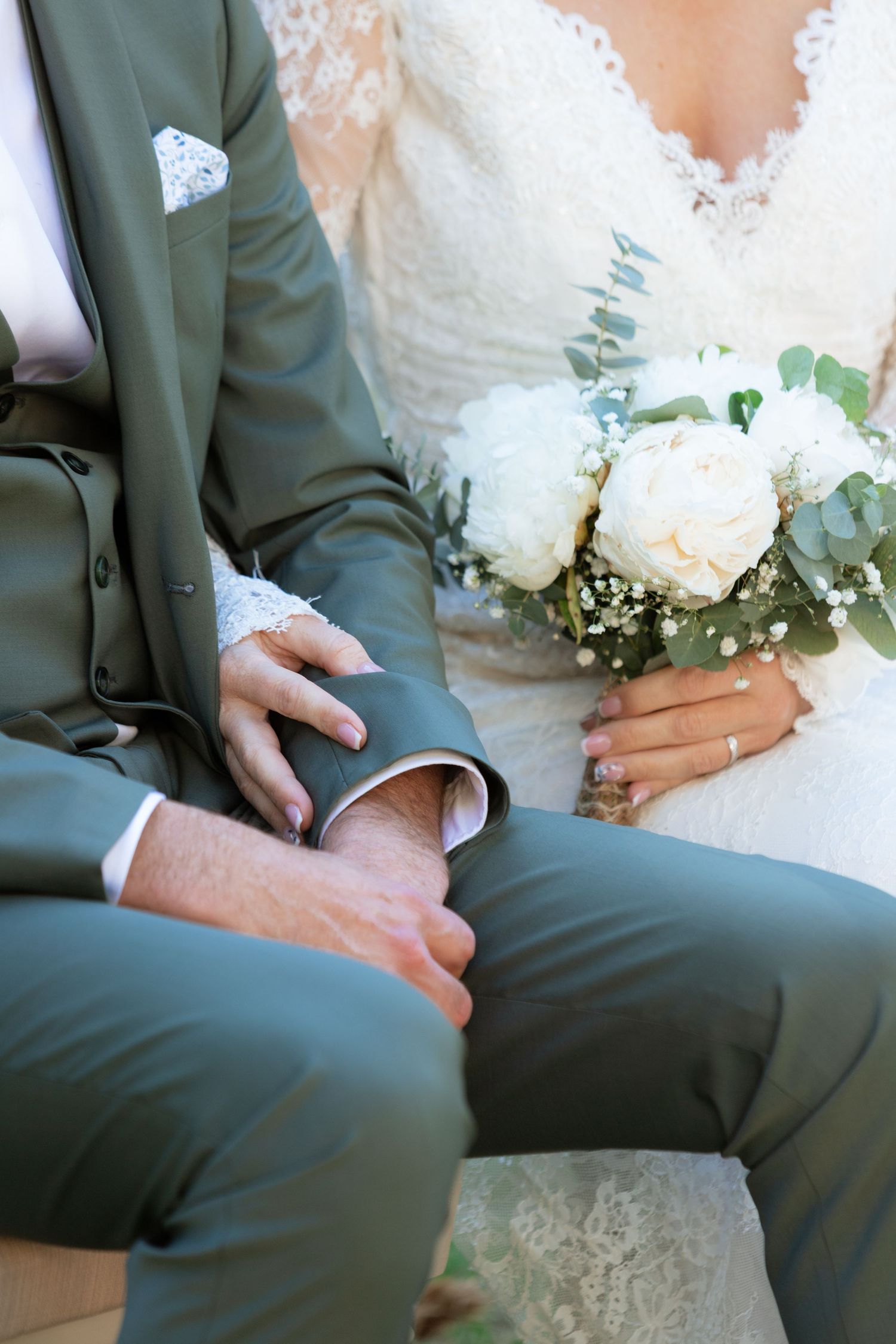 Bride and groom sitting together, holding hands, with a white peony bouquet and eucalyptus, wearing elegant wedding attire.