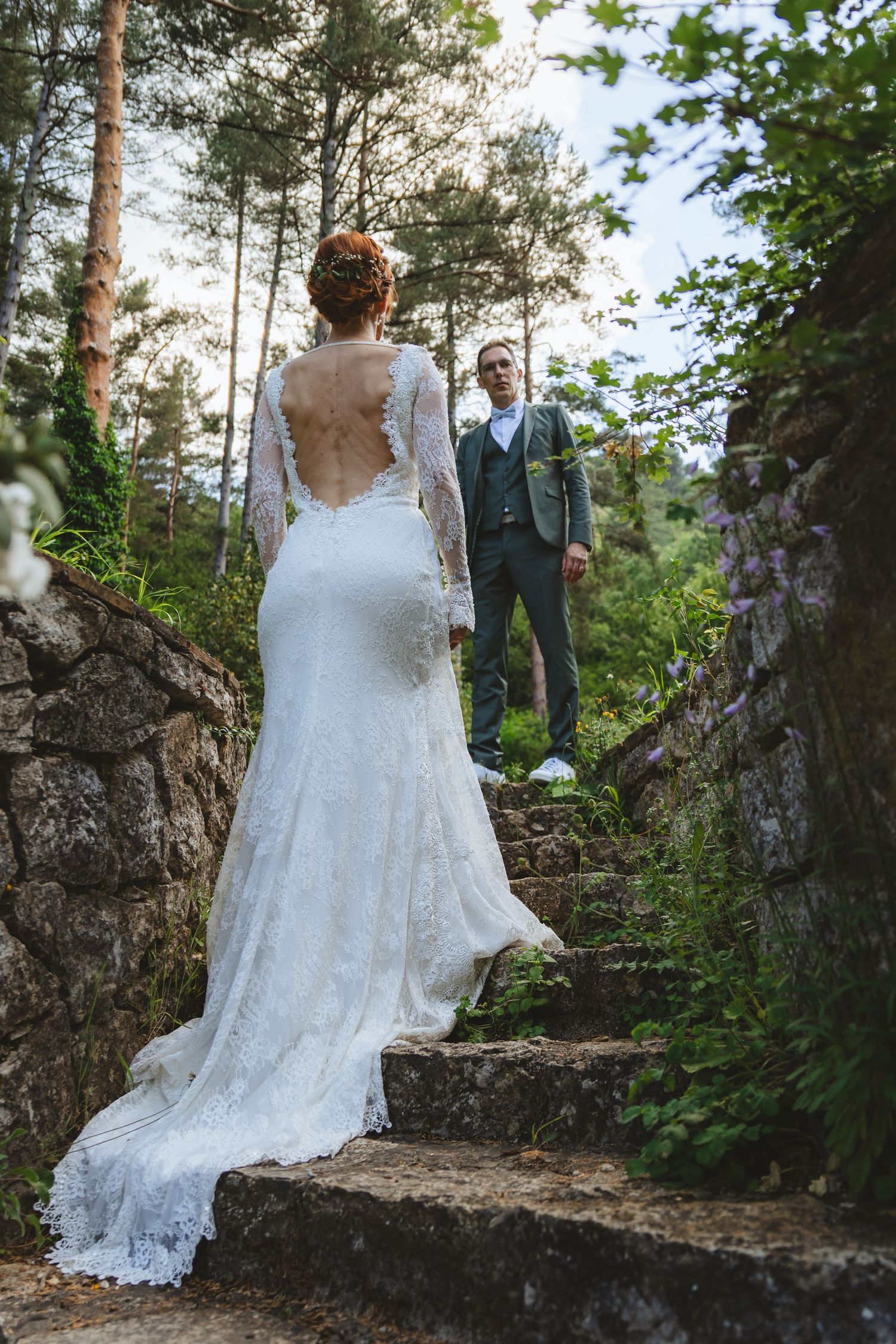 Bride in lace backless gown walks toward groom in gray suit on stone steps surrounded by lush forest trees.