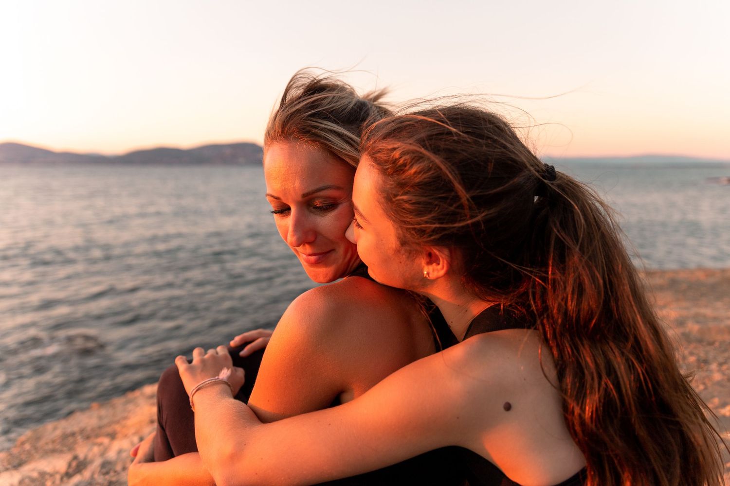 Two women sharing a warm embrace by the ocean at sunset, with wind-blown hair and a serene coastal backdrop.