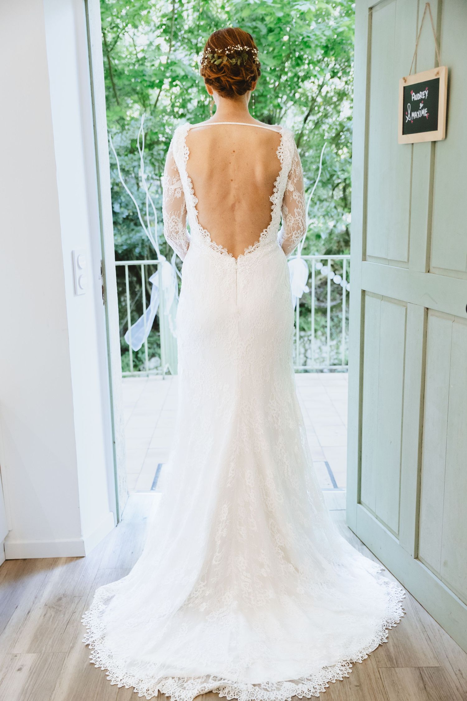 Bride in elegant backless lace wedding gown with long sleeves and train, standing in doorway overlooking garden.