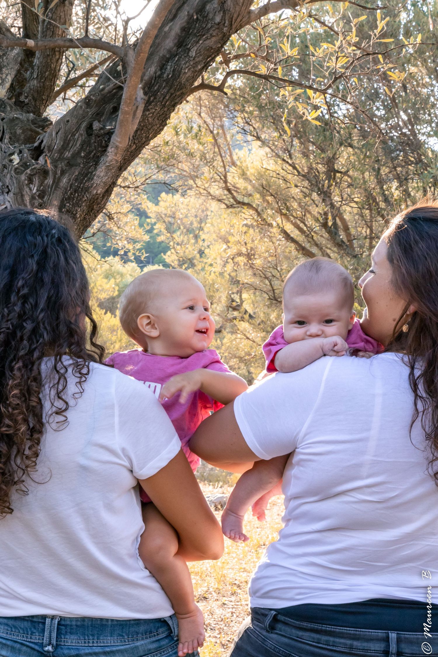 Two mothers in white shirts hold their babies face-to-face under a tree outdoors.