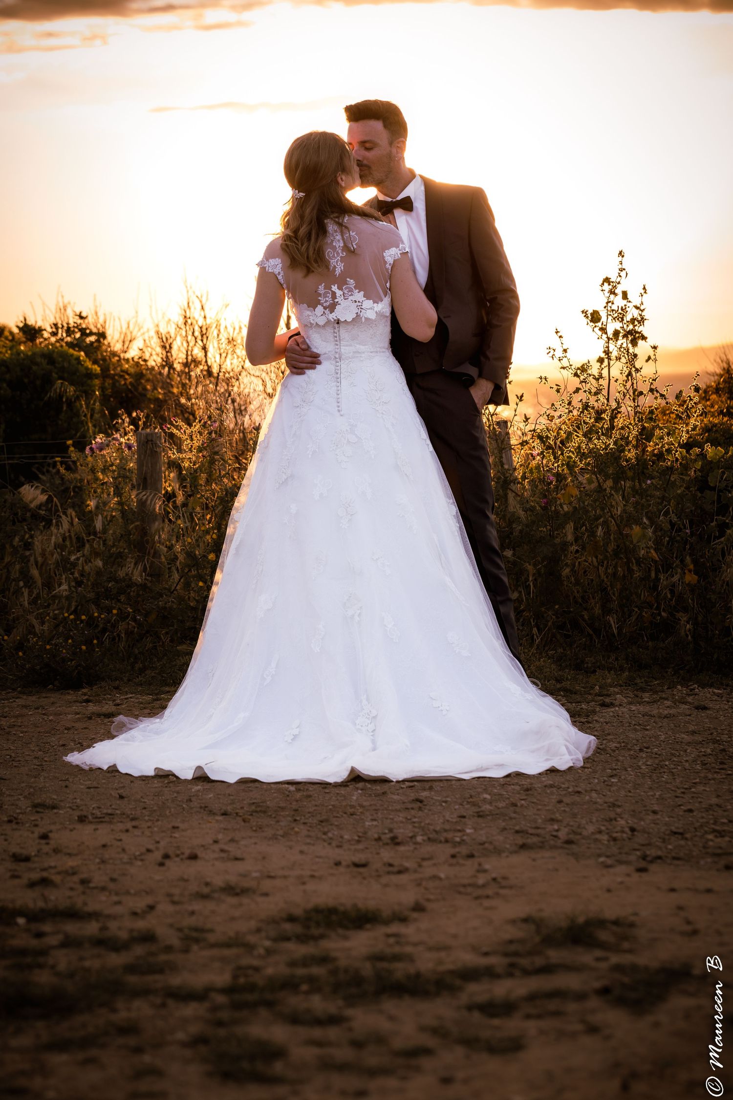 Bride and groom kissing at sunset in a field, bride wearing elegant lace wedding gown with train.