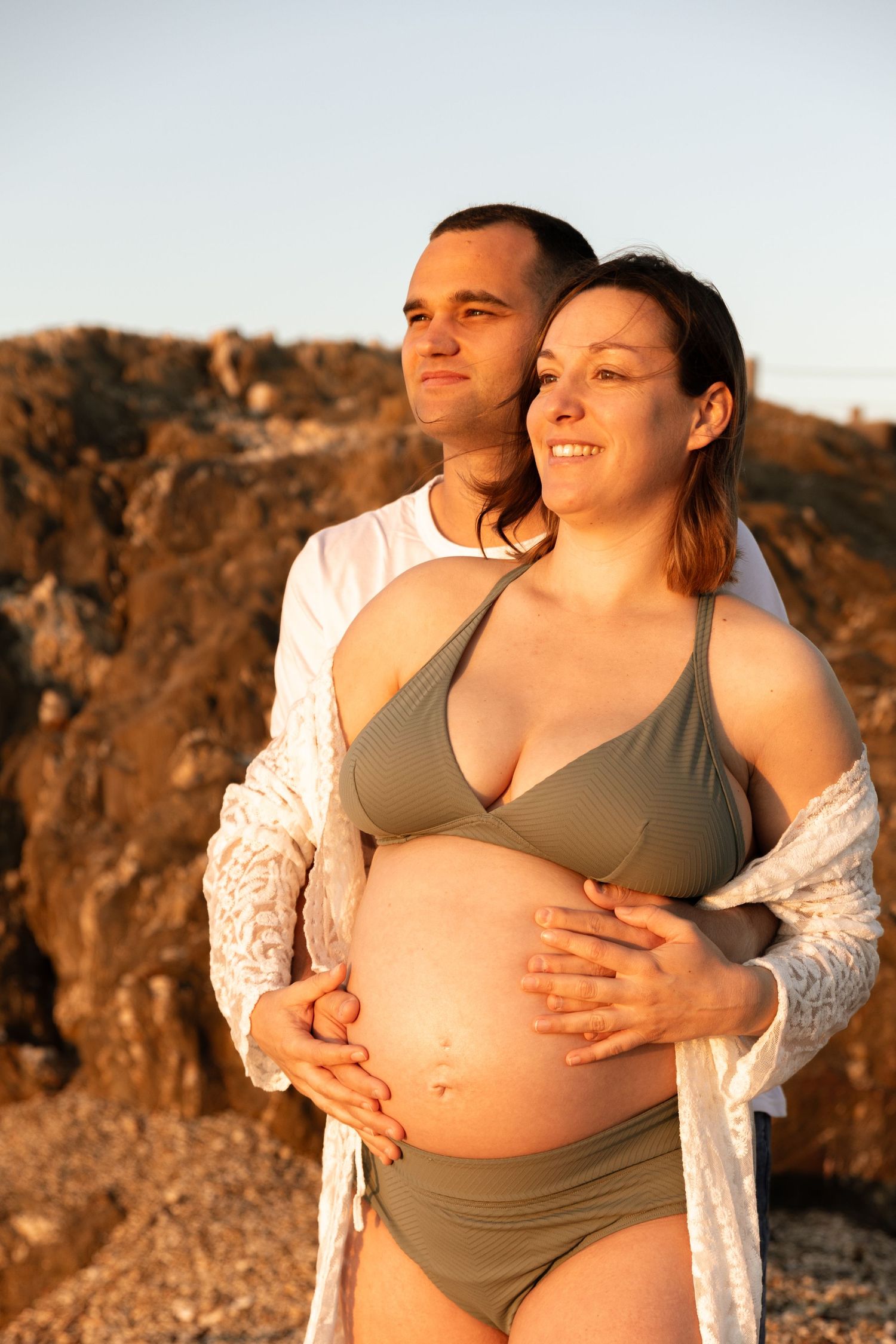 Happy pregnant woman in bikini embraced by partner against rocky coastal backdrop in warm sunset light.