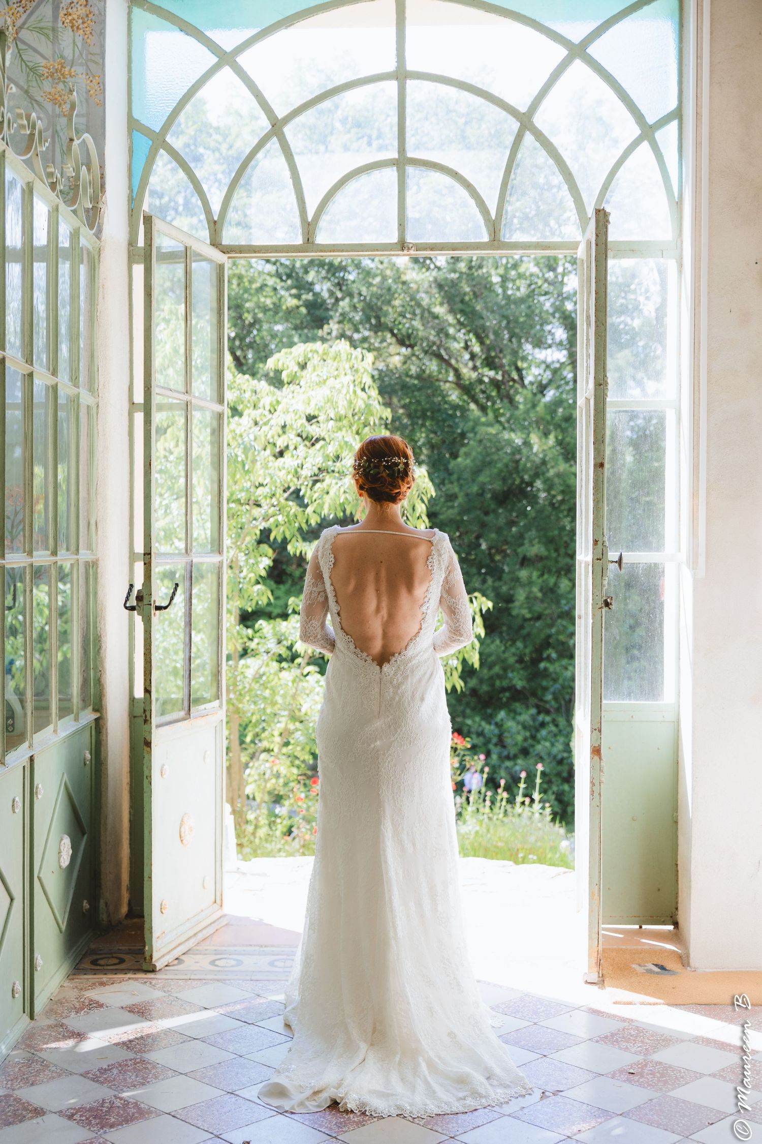 Bride in backless lace wedding gown standing in arched glass doors overlooking a lush garden.