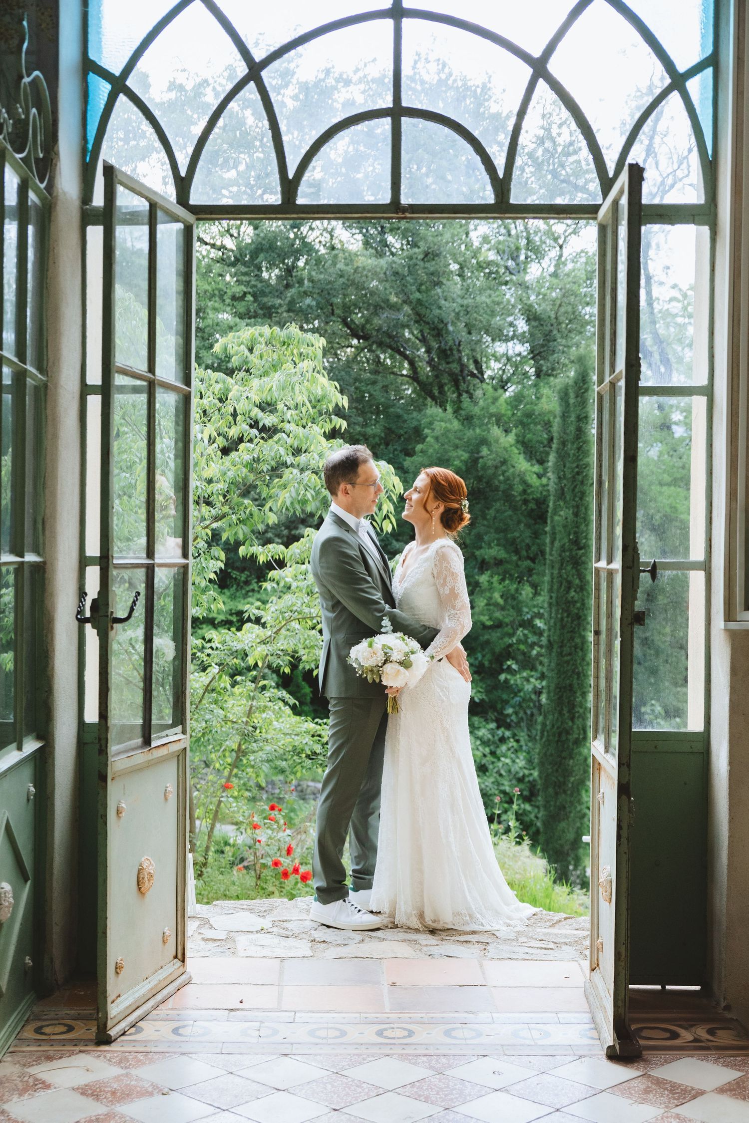 Bride and groom embrace in elegant arched glass doorway, lush green garden backdrop, white bouquet, wedding attire.