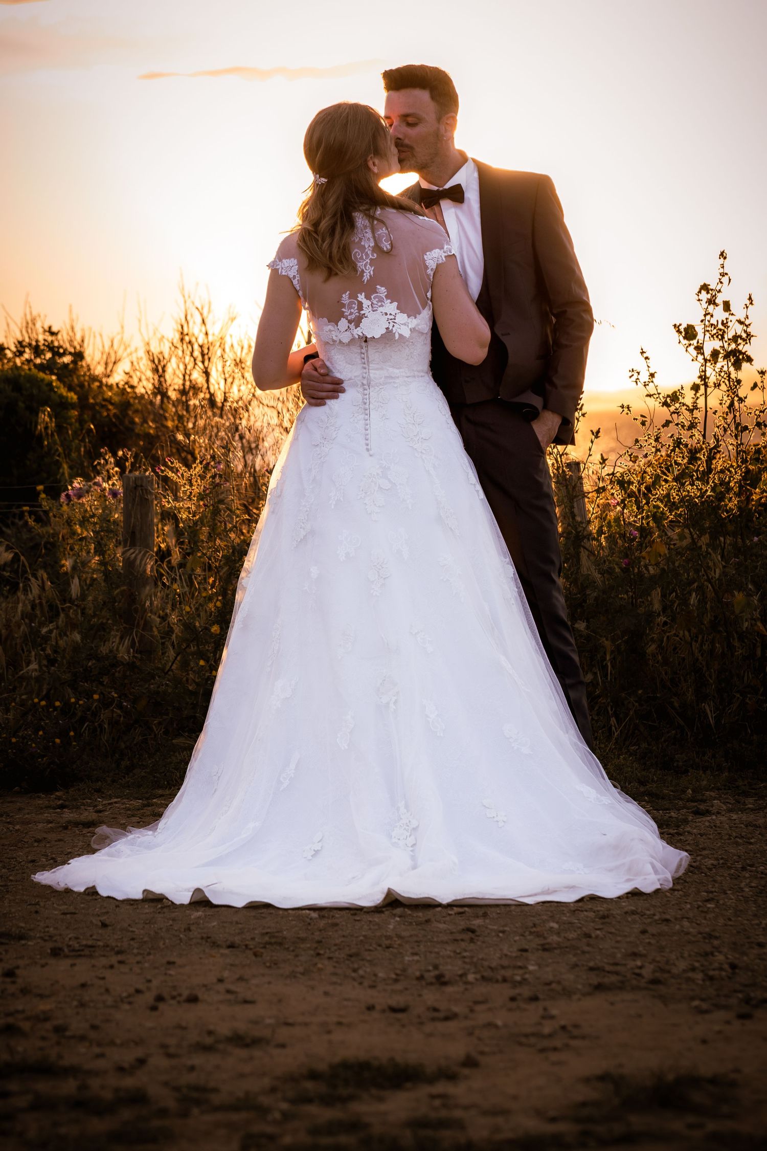 Bride and groom sharing a romantic kiss at sunset, with bride in elegant lace wedding gown and groom in dark suit.