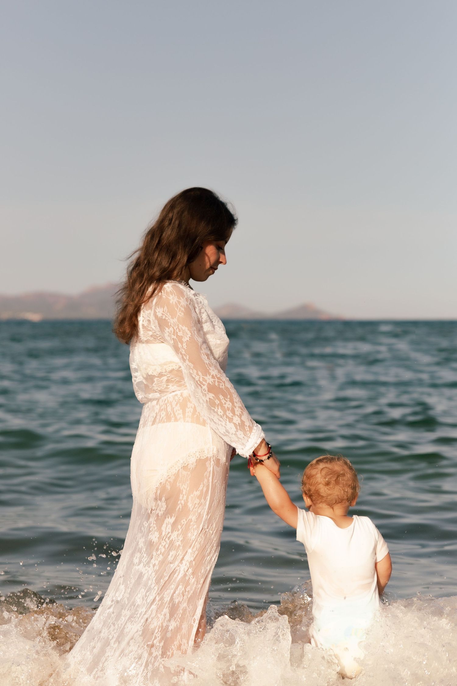 Pregnant woman in white lace dress holds toddler's hand while wading in ocean waves on a sunny day.