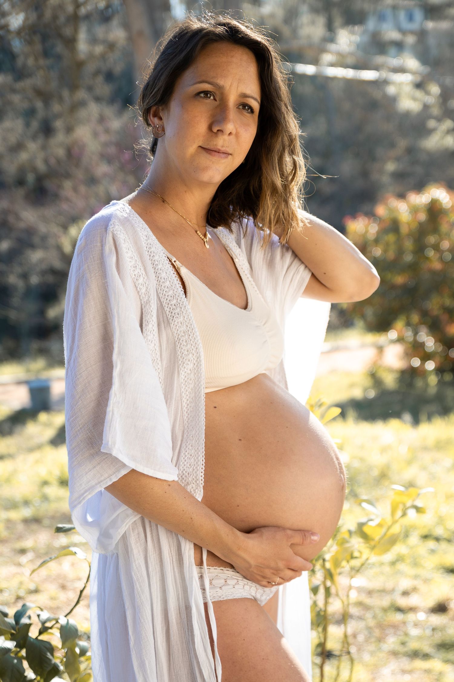 Pregnant woman in white robe and crop top holding her bare belly in a sunlit garden.