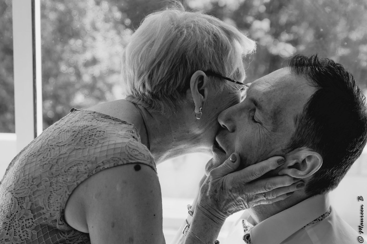 Elderly woman tenderly holds a man's face for a kiss in a black and white wedding photo.