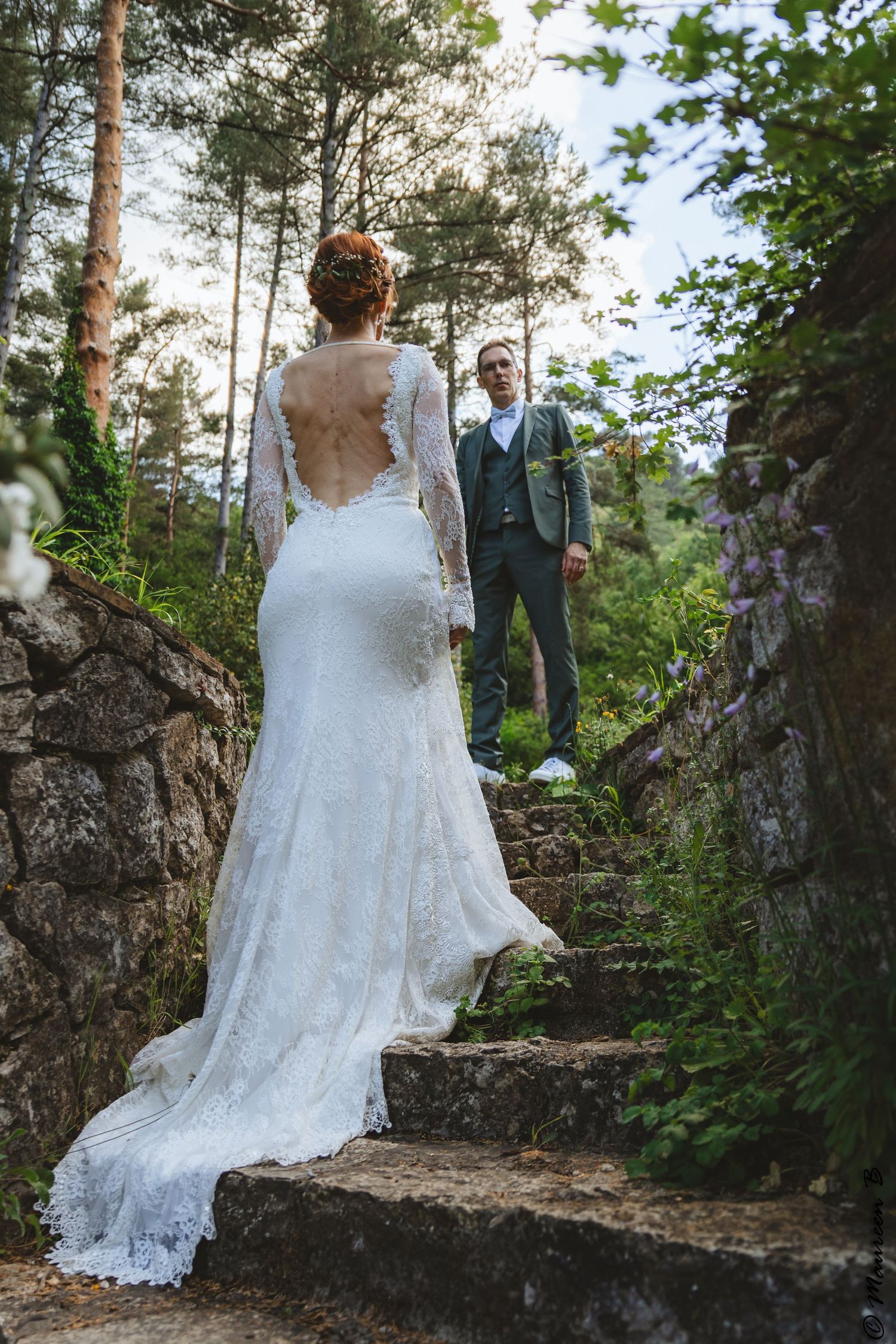 Bride in lace backless wedding gown walks toward groom on stone steps in a forest setting.