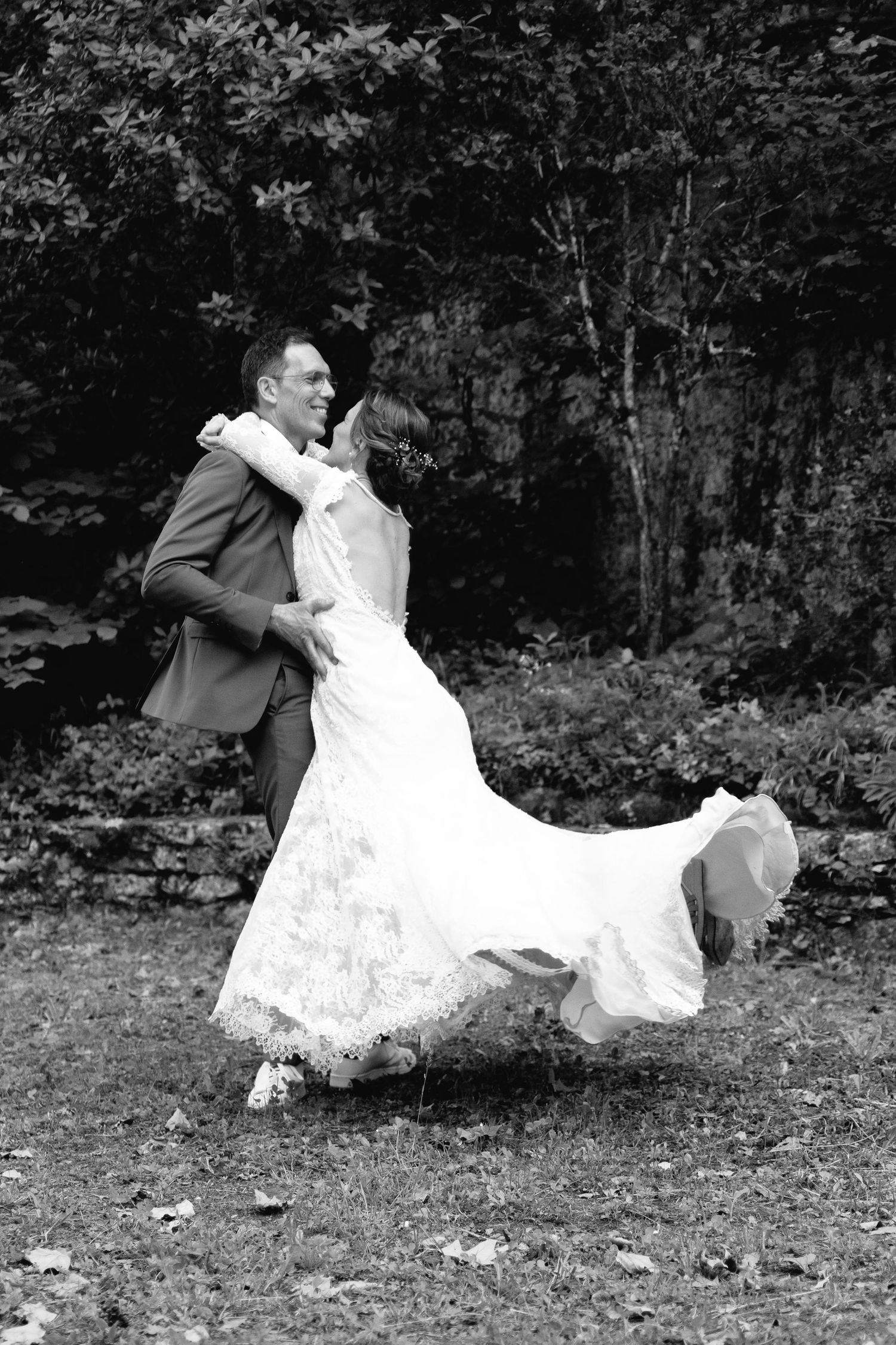 Black and white photo of a bride and groom dancing joyfully outdoors, her lace dress swirling, surrounded by lush trees.