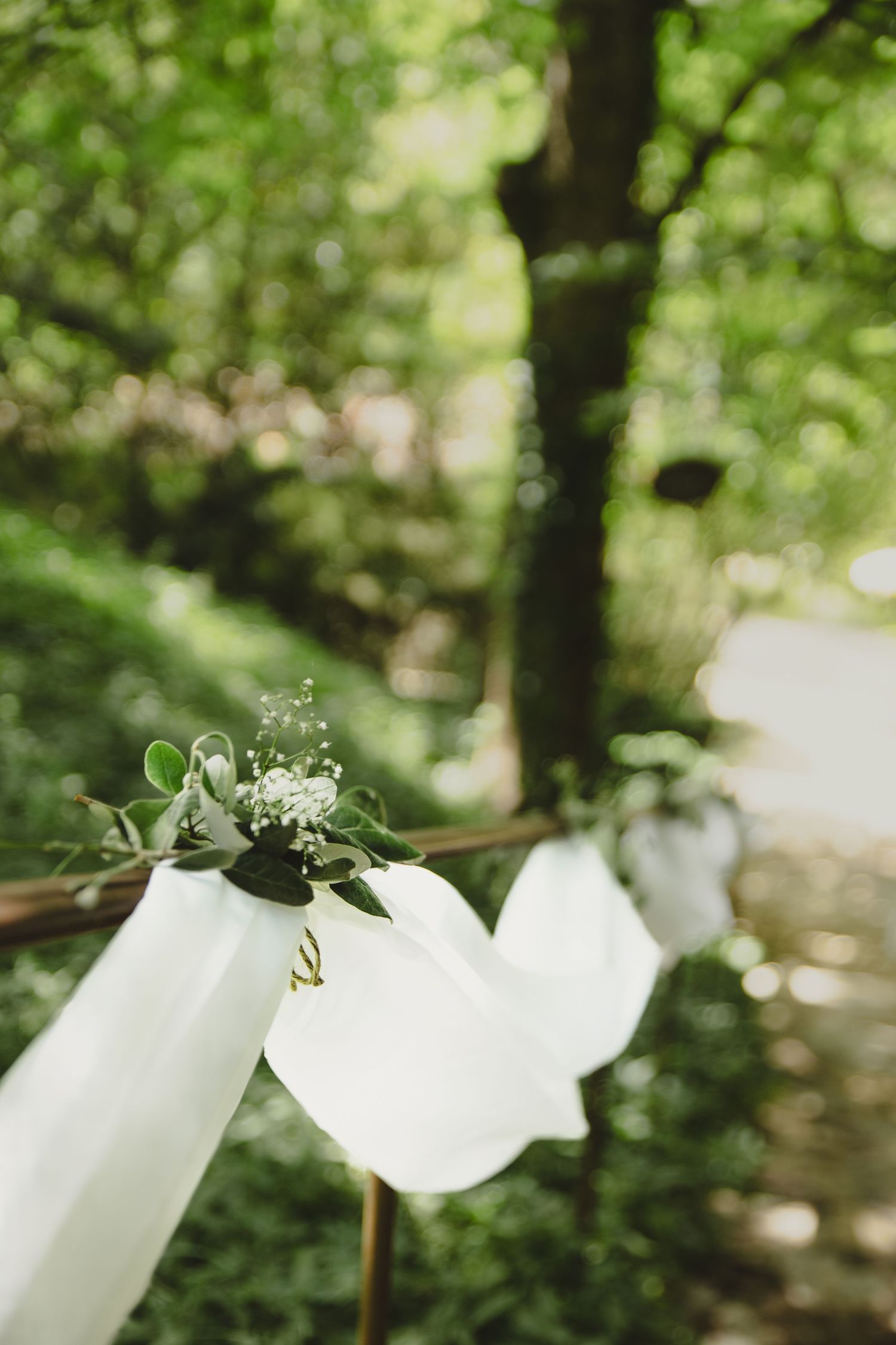 White fabric and floral garland draped on wooden poles in a lush green forest wedding ceremony setting.
