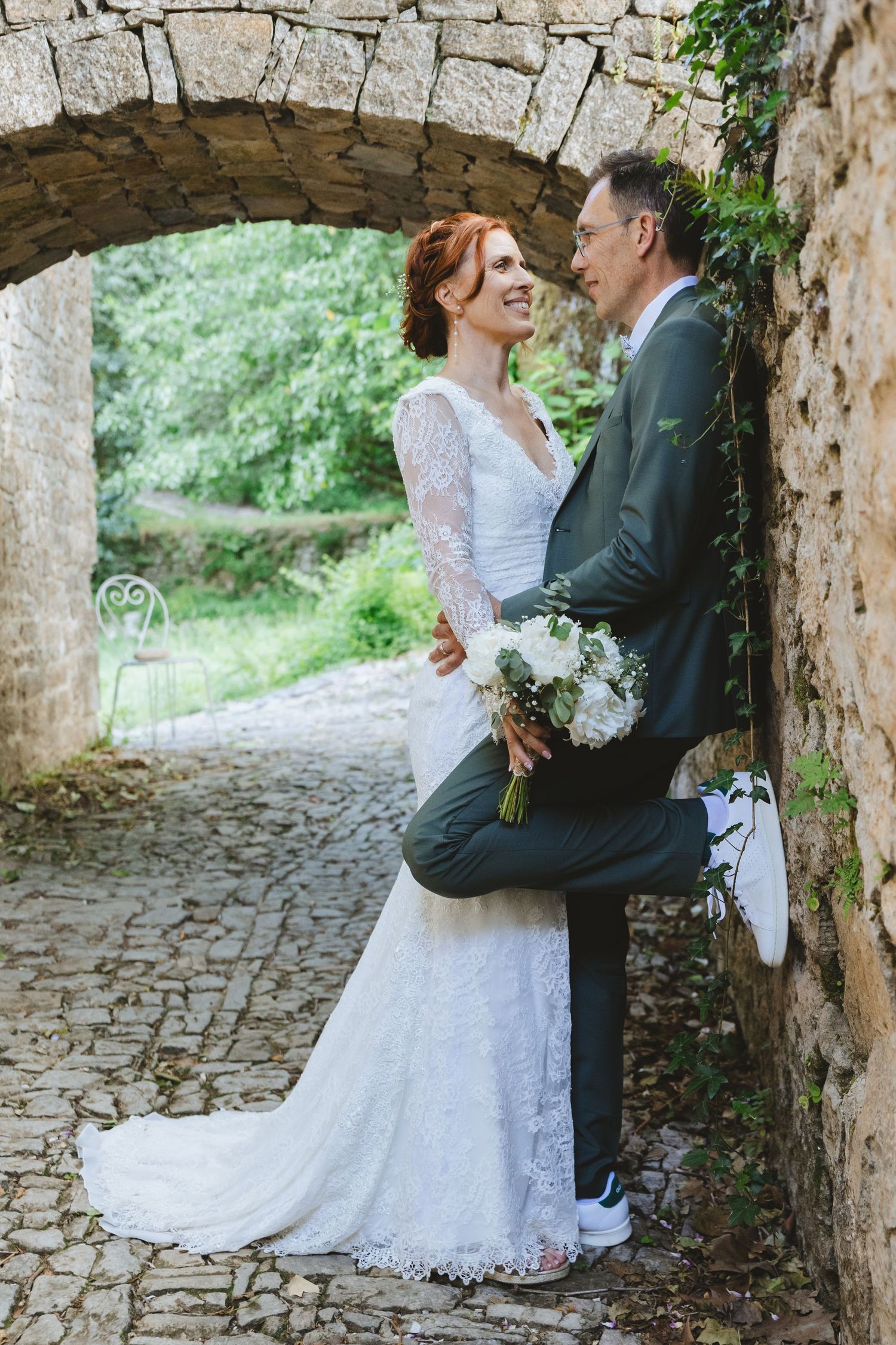 Bride in lace gown and groom in gray suit share a romantic moment under a stone arch with a white bouquet.
