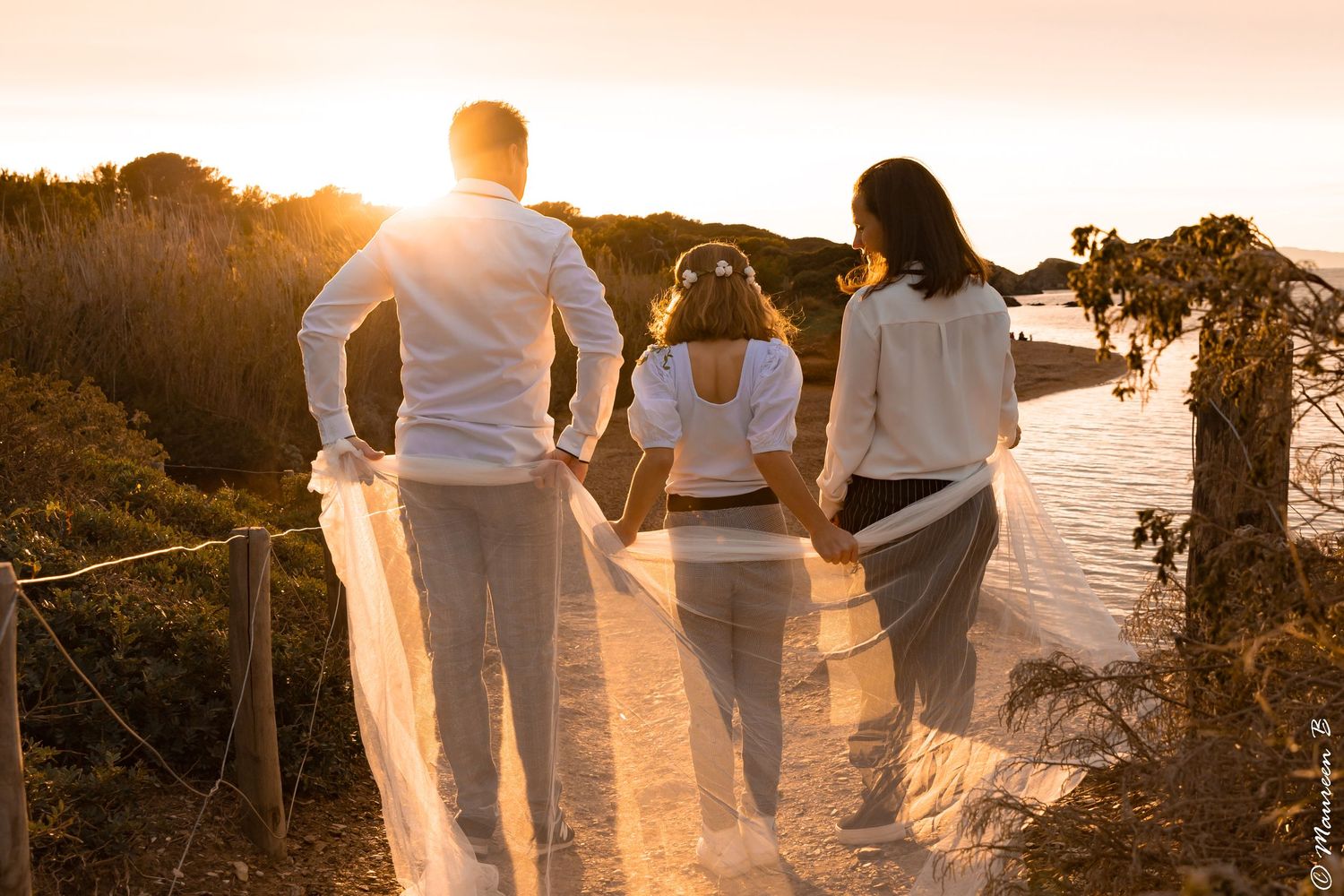 Family of three holding hands watching sunset over ocean from coastal path.