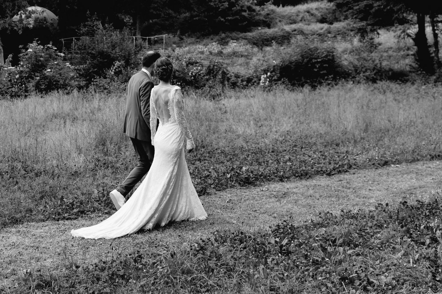 Black and white photo of a bride and groom walking together through a lush green field on their wedding day.