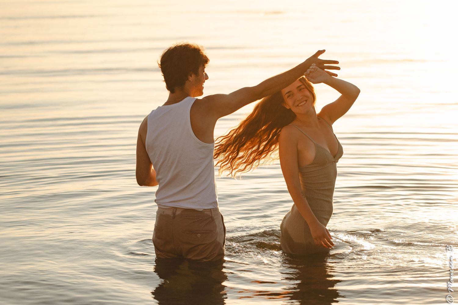 Couple dancing playfully in the ocean at golden sunset, woman tossing her long hair back.