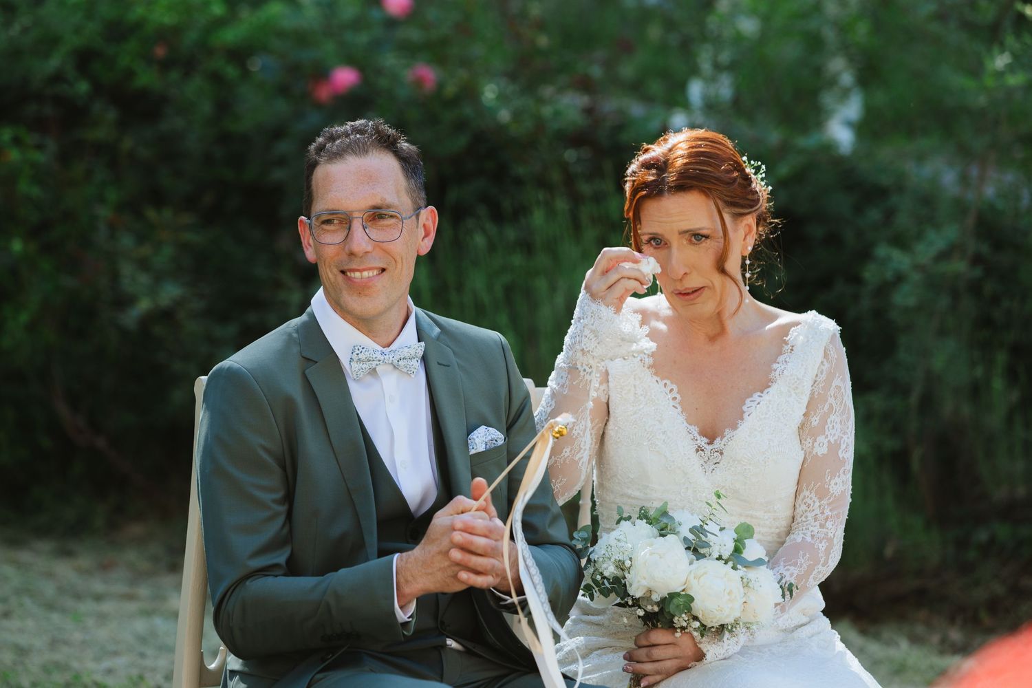 Bride wiping tears while groom holds champagne bottle during outdoor wedding ceremony surrounded by lush greenery.