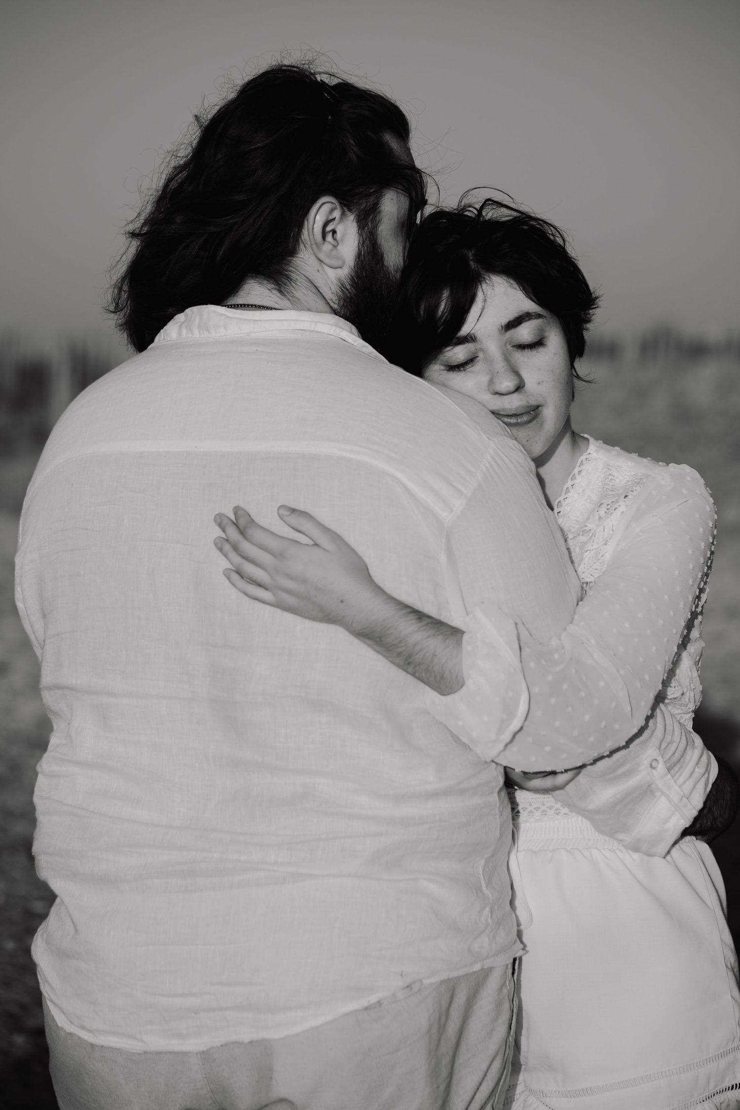 Black and white photo of a couple embracing tenderly outdoors, both wearing white linen clothing.
