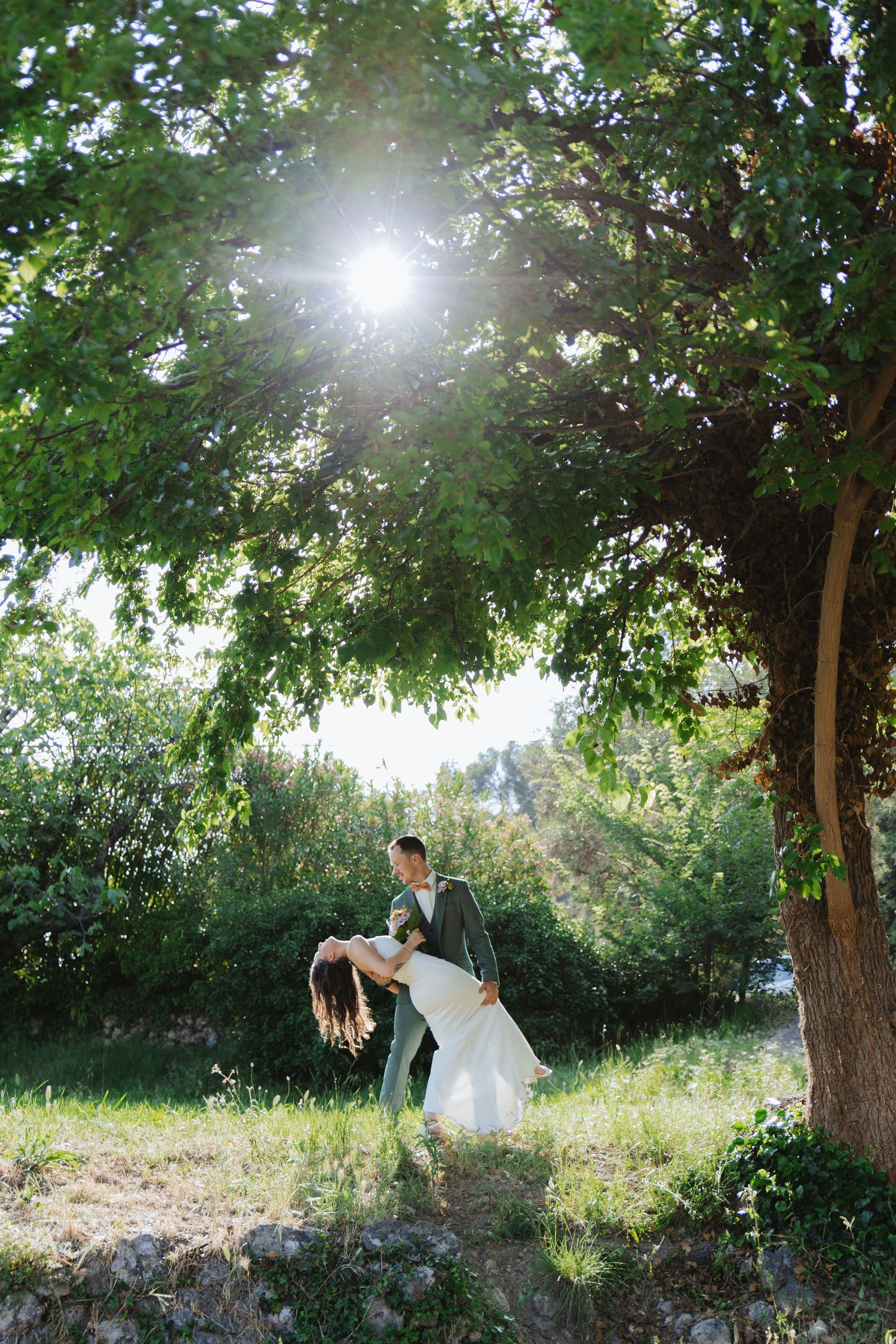 Groom dips bride under a sunlit tree in a lush green garden during an outdoor wedding ceremony.