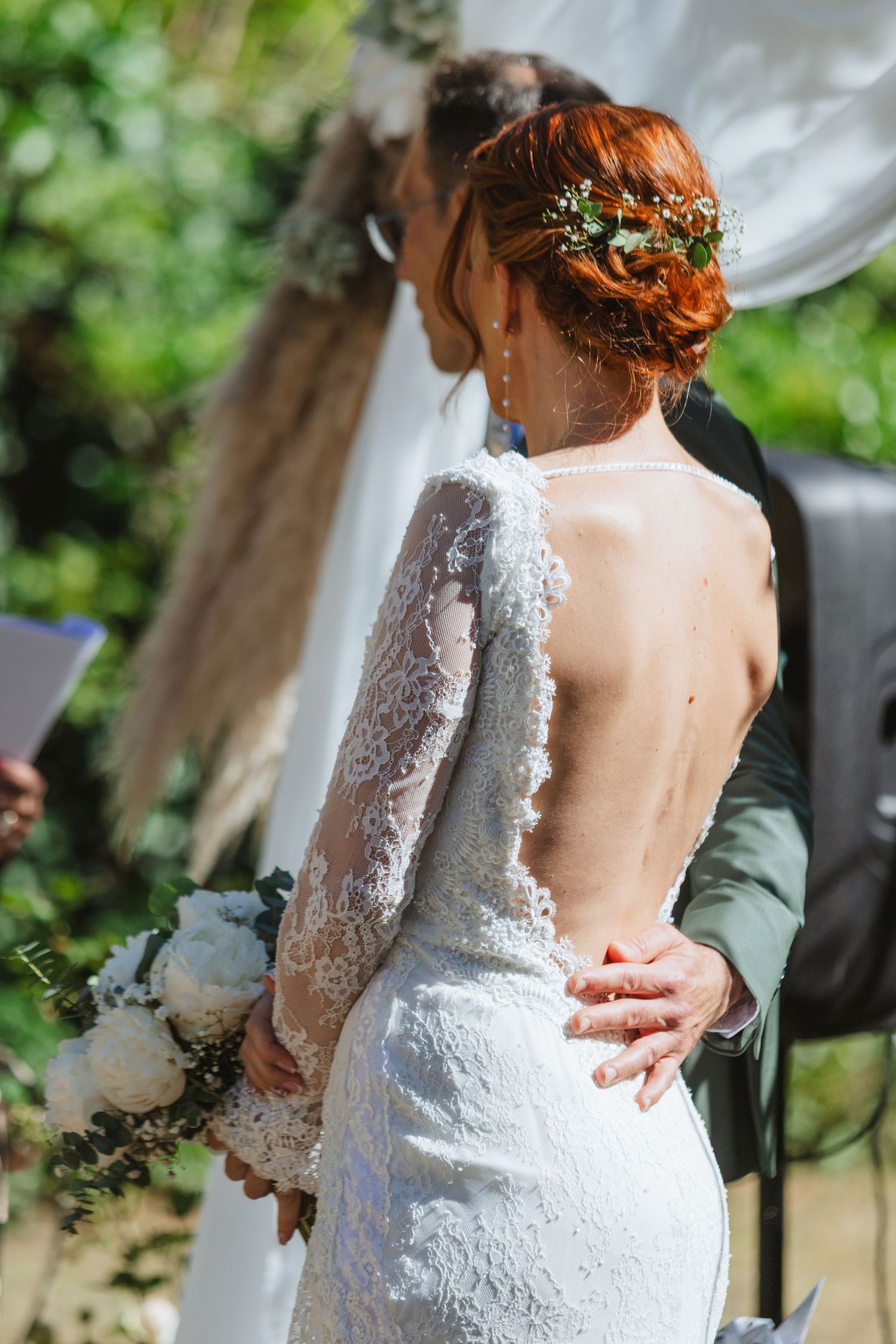 Bride in backless lace wedding dress with floral updo holds white bouquet during outdoor ceremony.