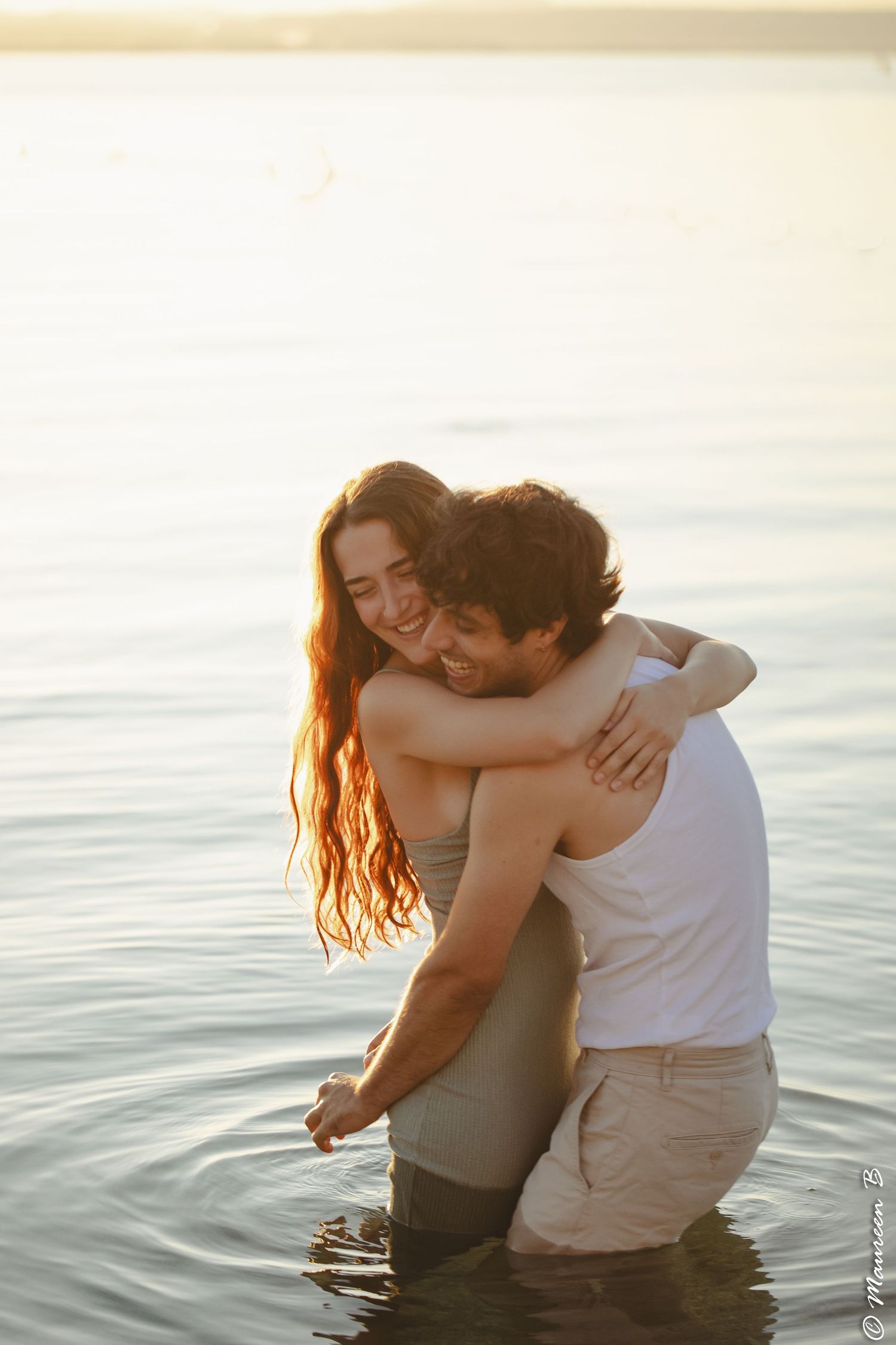 Joyful couple embracing and laughing together while standing in calm golden sunset lake waters.