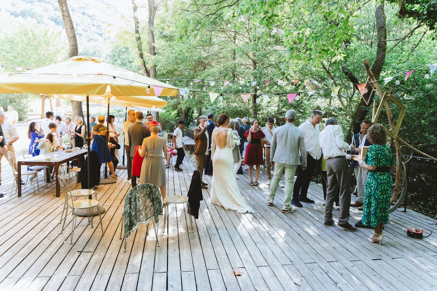 Outdoor wedding reception on a wooden deck surrounded by lush green trees with guests mingling in elegant attire.
