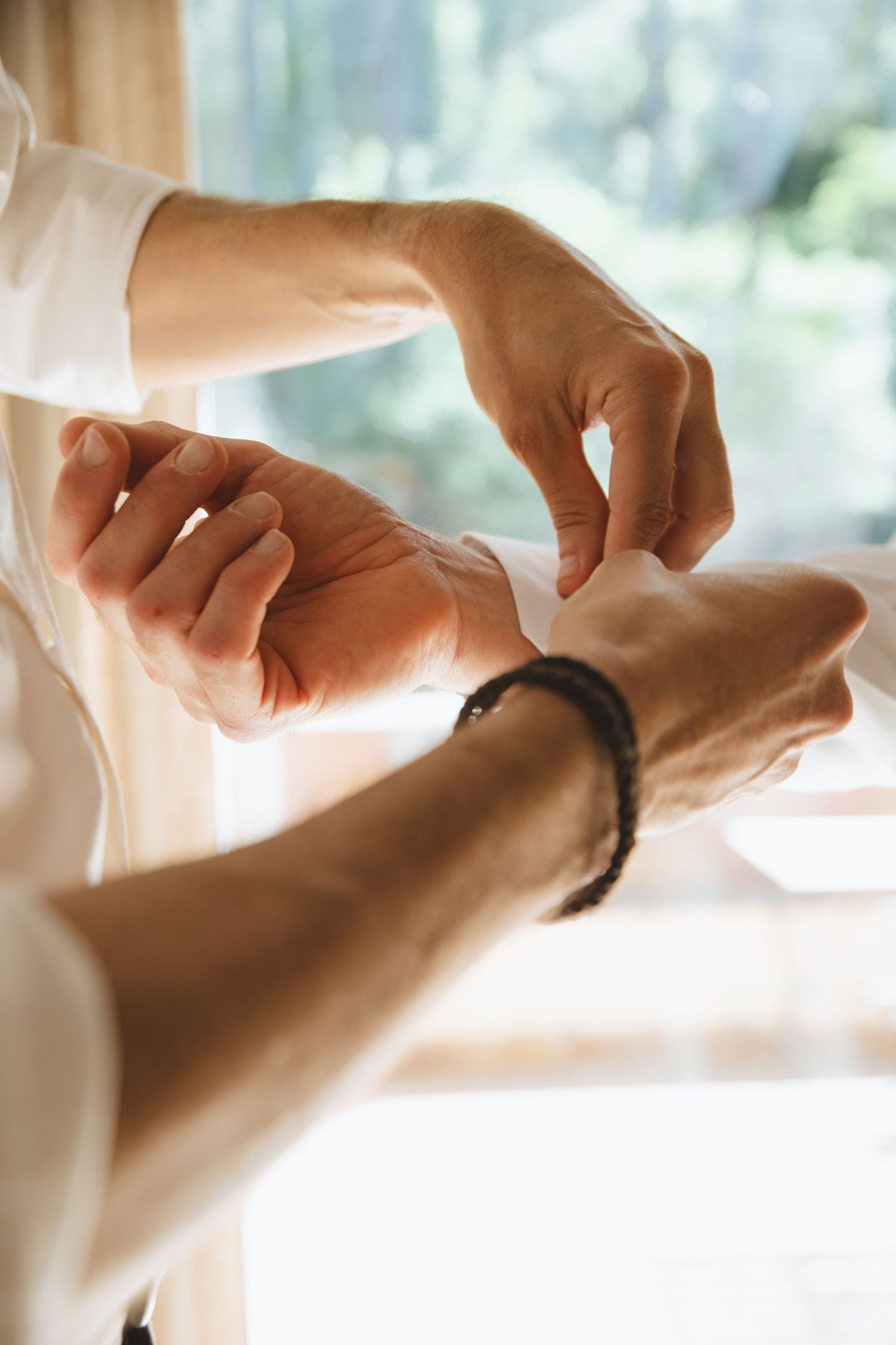 Man fastening white dress shirt cuff with a braided bracelet on his wrist near a bright window.