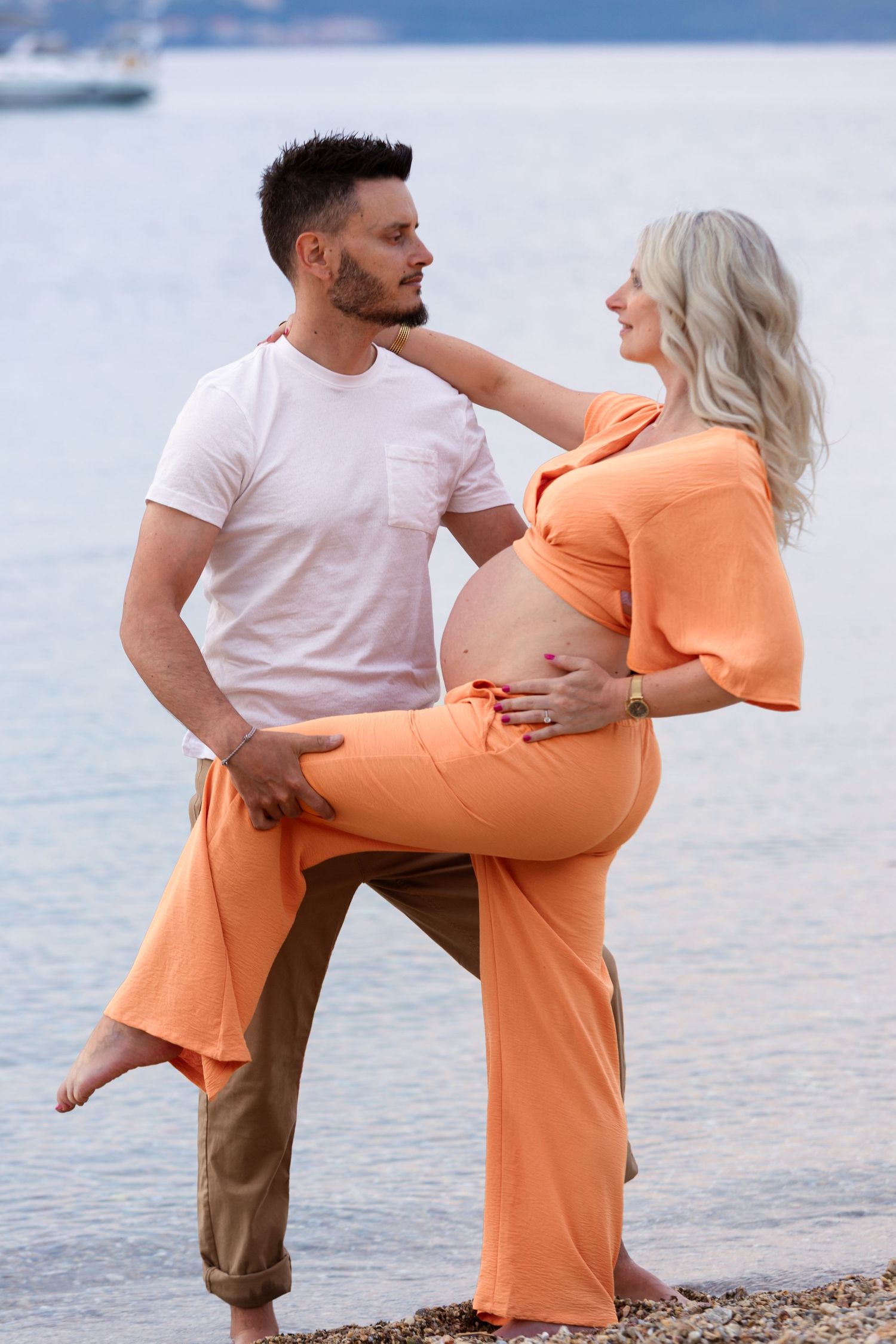 Couple sharing a tender moment on a rocky beach, pregnant woman in orange outfit with partner in white shirt near calm water.