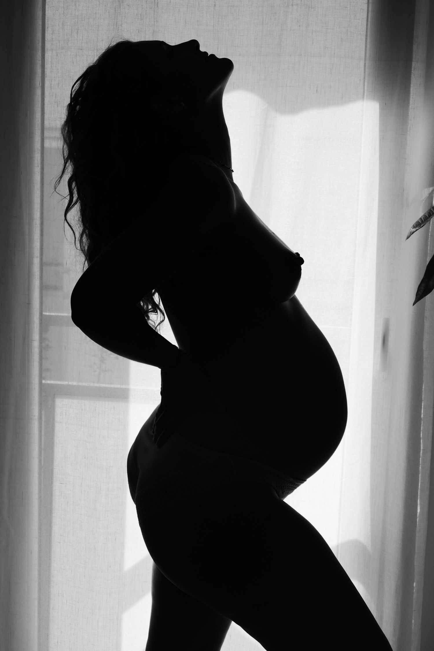 Black and white silhouette of a pregnant woman standing by a window, showcasing her baby bump.