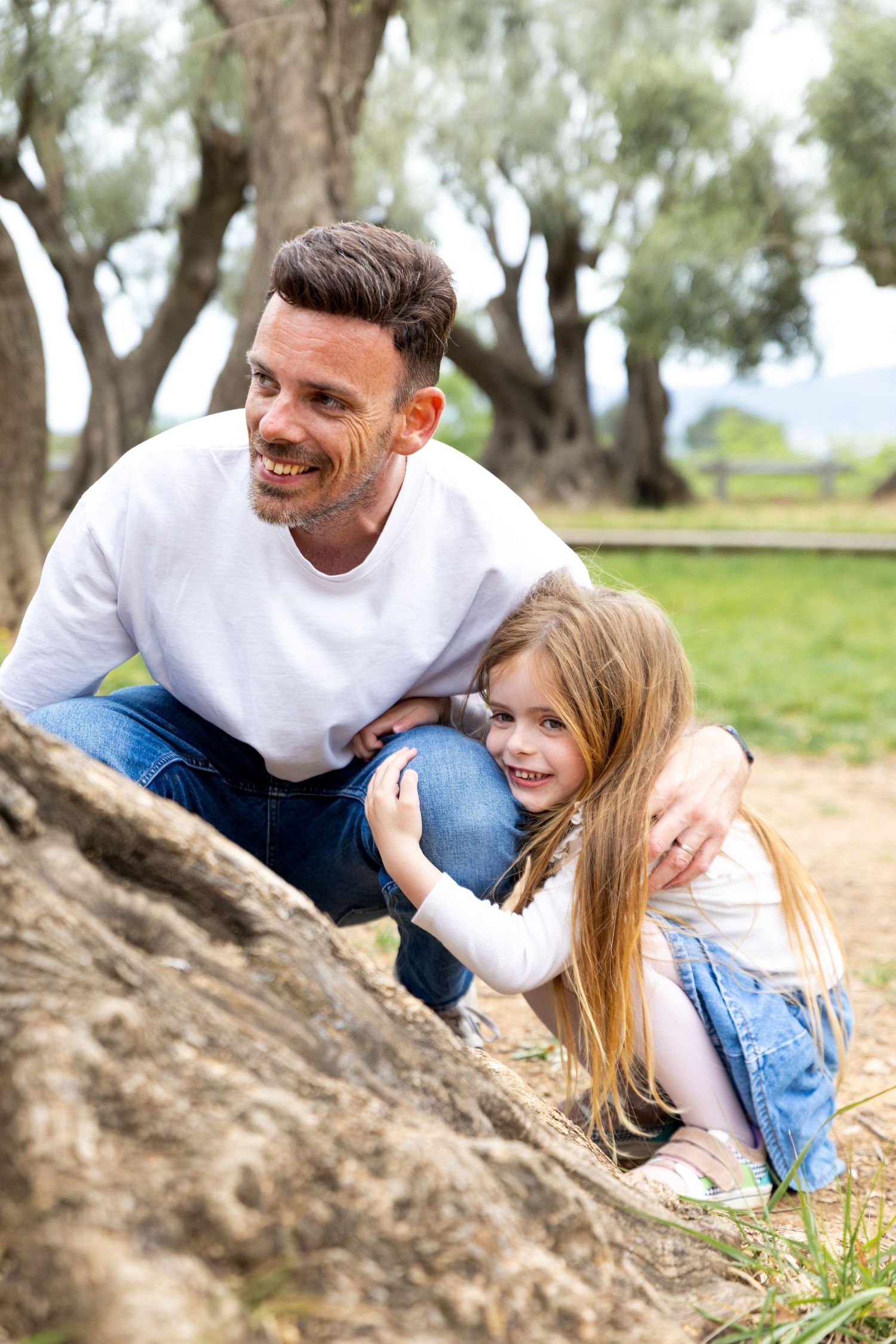 A smiling father crouches beside his young daughter near a tree trunk in an olive grove.