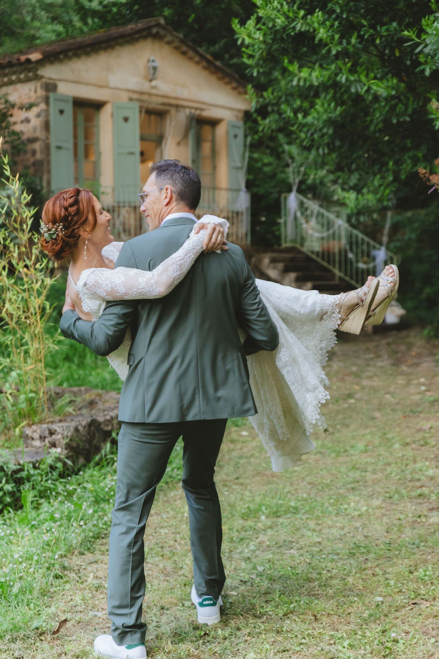 Groom in gray suit carries bride in lace wedding dress outdoors near rustic stone building surrounded by lush greenery.