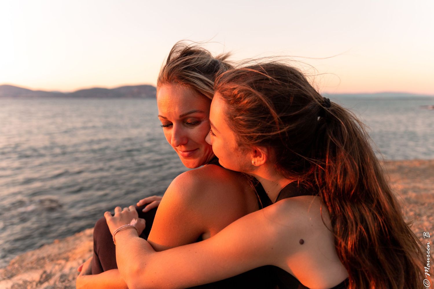 Two women share a warm embrace on a rocky beach during a beautiful golden sunset.