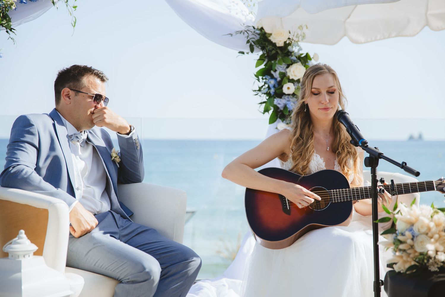A bride plays acoustic guitar at a beachside wedding ceremony while the groom sits beside her under a floral arch.