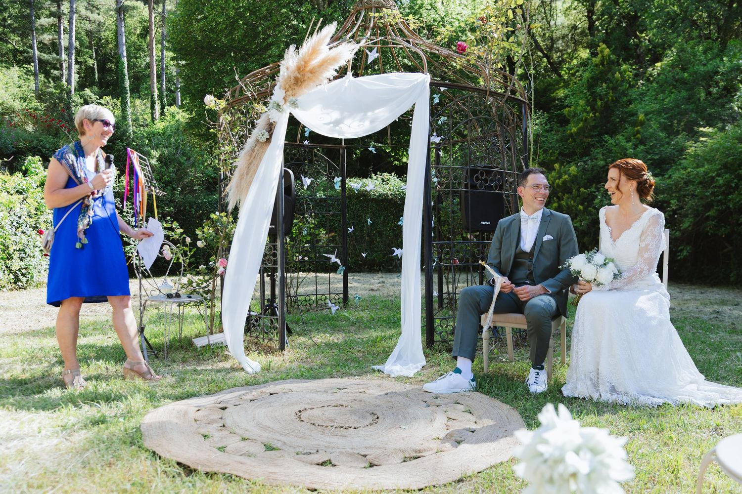 Outdoor wedding ceremony with bride, groom seated and officiant in blue dress under decorated arch in garden.