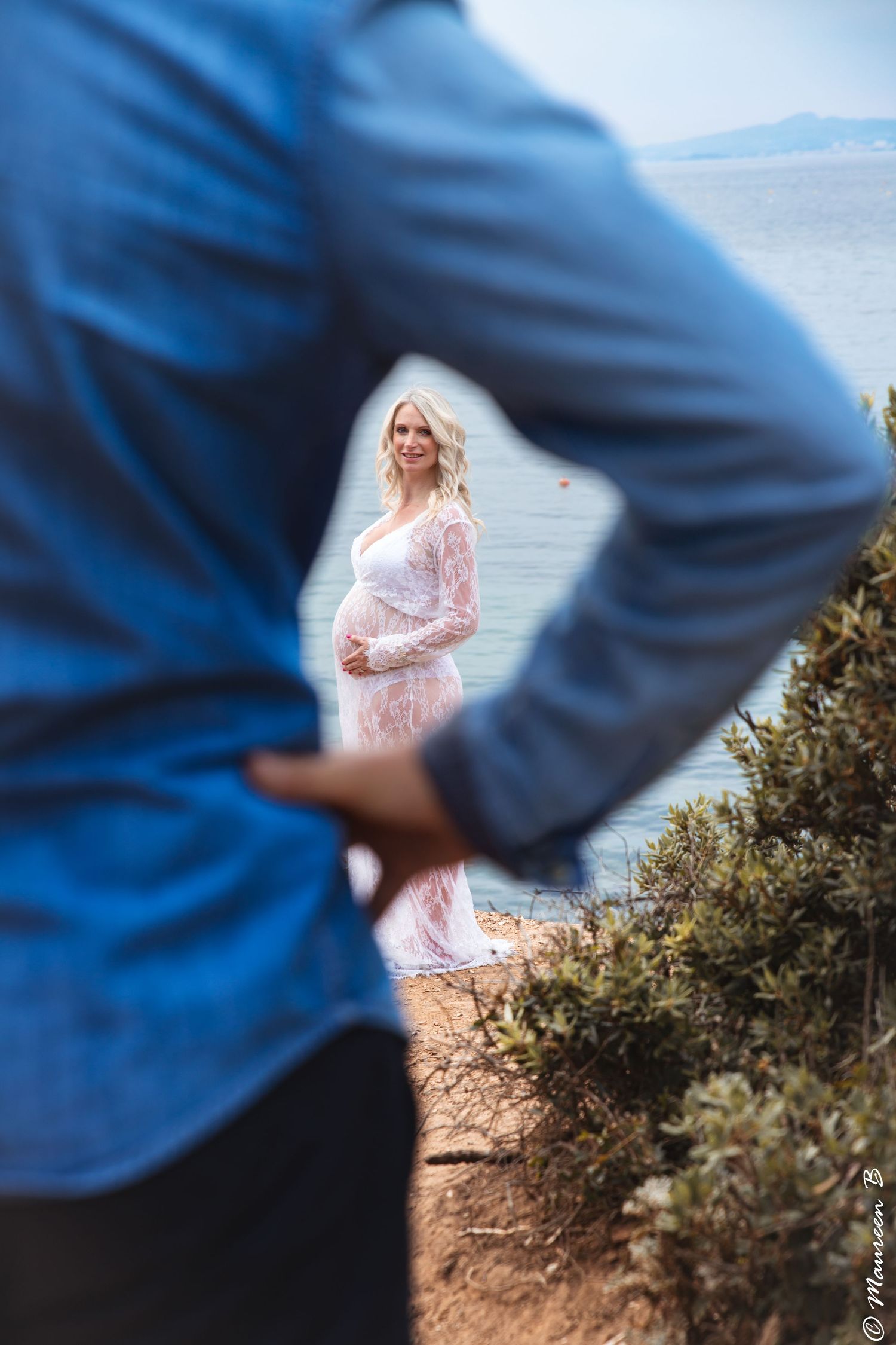 Creative maternity photo framing pregnant blonde woman in white lace dress through partner's arms outdoors.