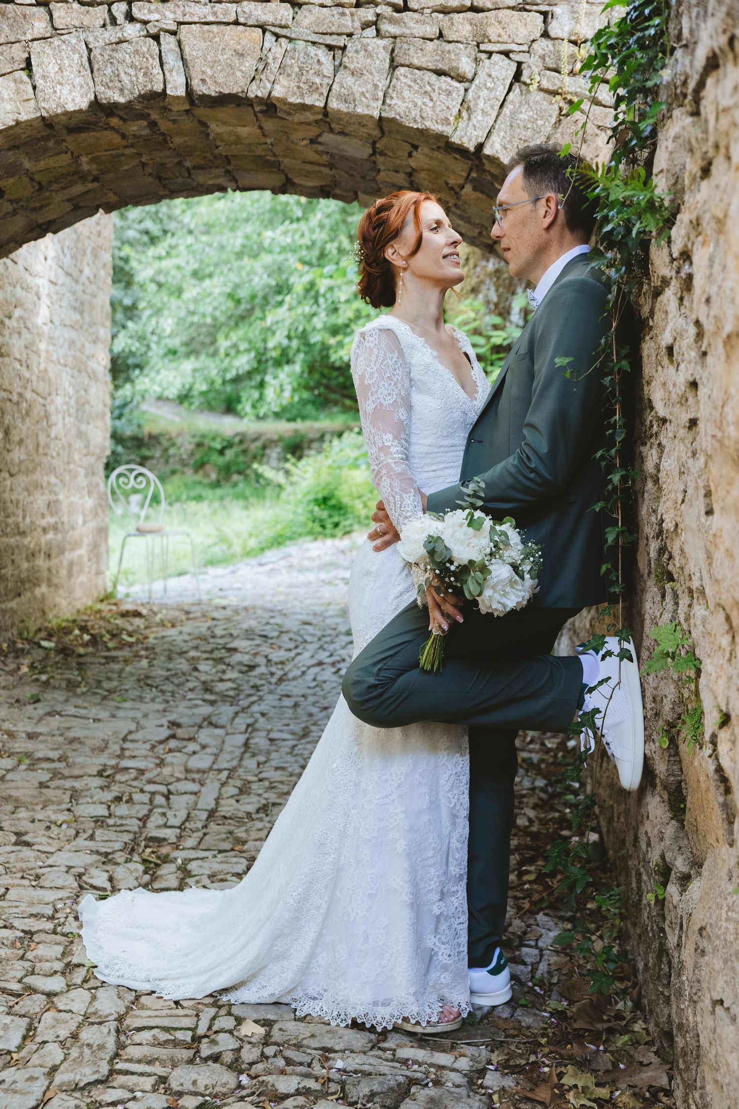 Bride and groom share a romantic moment under a stone arch, she holds white flowers, wearing lace gown, he in gray suit.