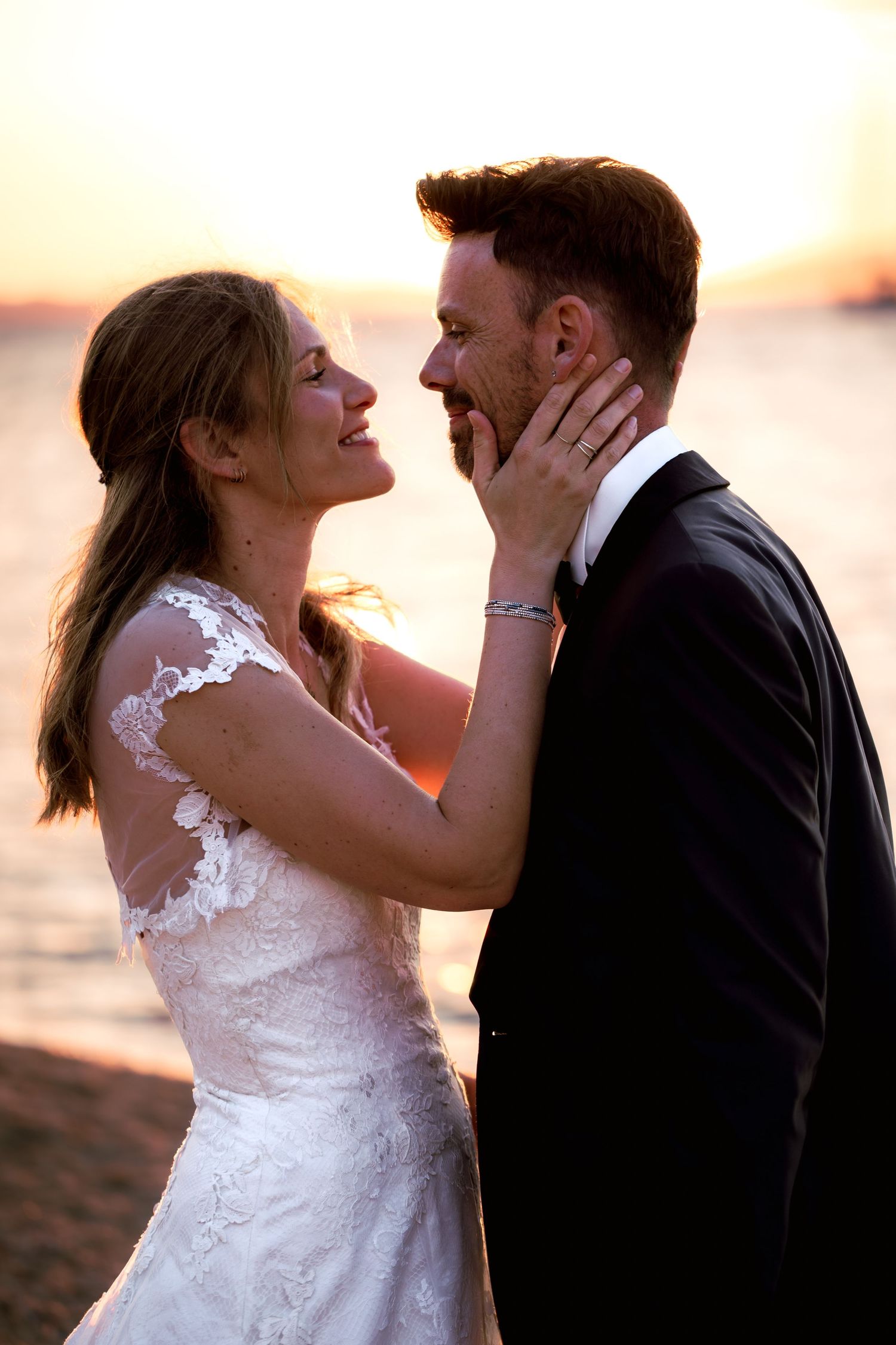 Bride and groom share a romantic sunset moment by the water, smiling at each other in wedding attire.