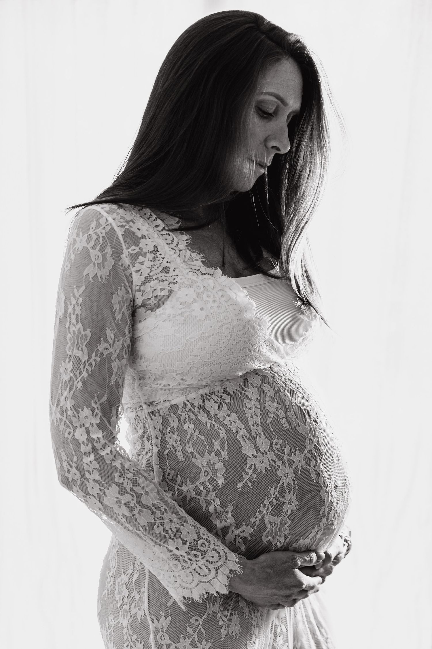 Pregnant woman in white lace dress cradling baby bump in black and white portrait.