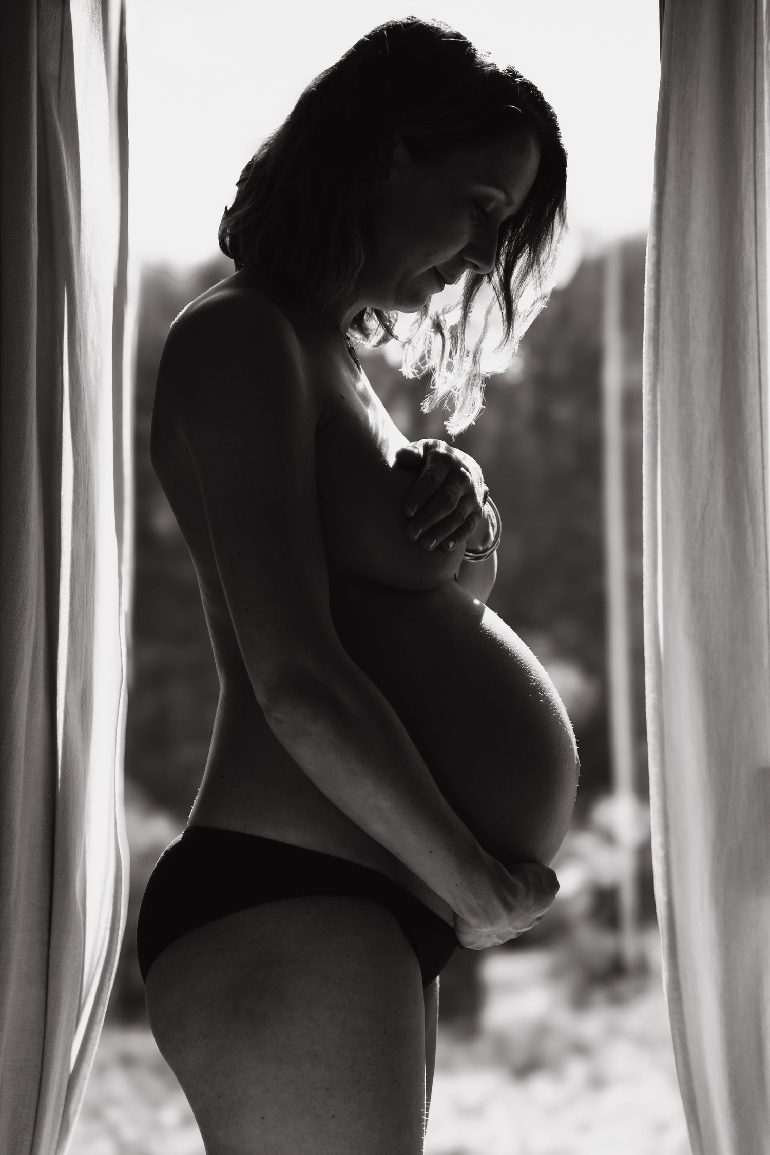 Black and white silhouette of a pregnant woman standing by a window, cradling her baby bump.