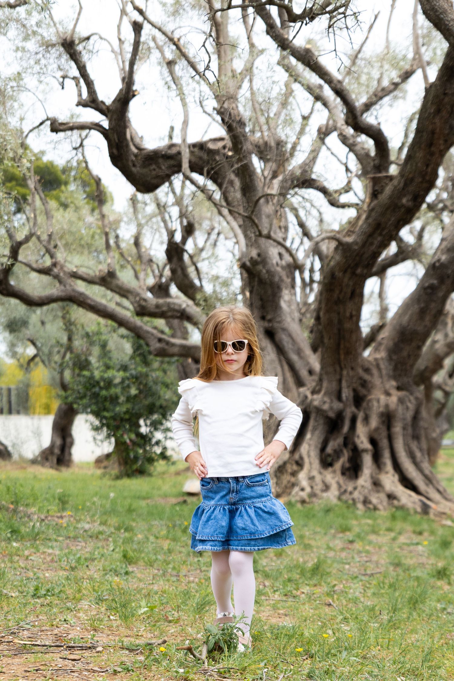 Young girl in white top and denim skirt poses confidently in sunglasses before a large tree.