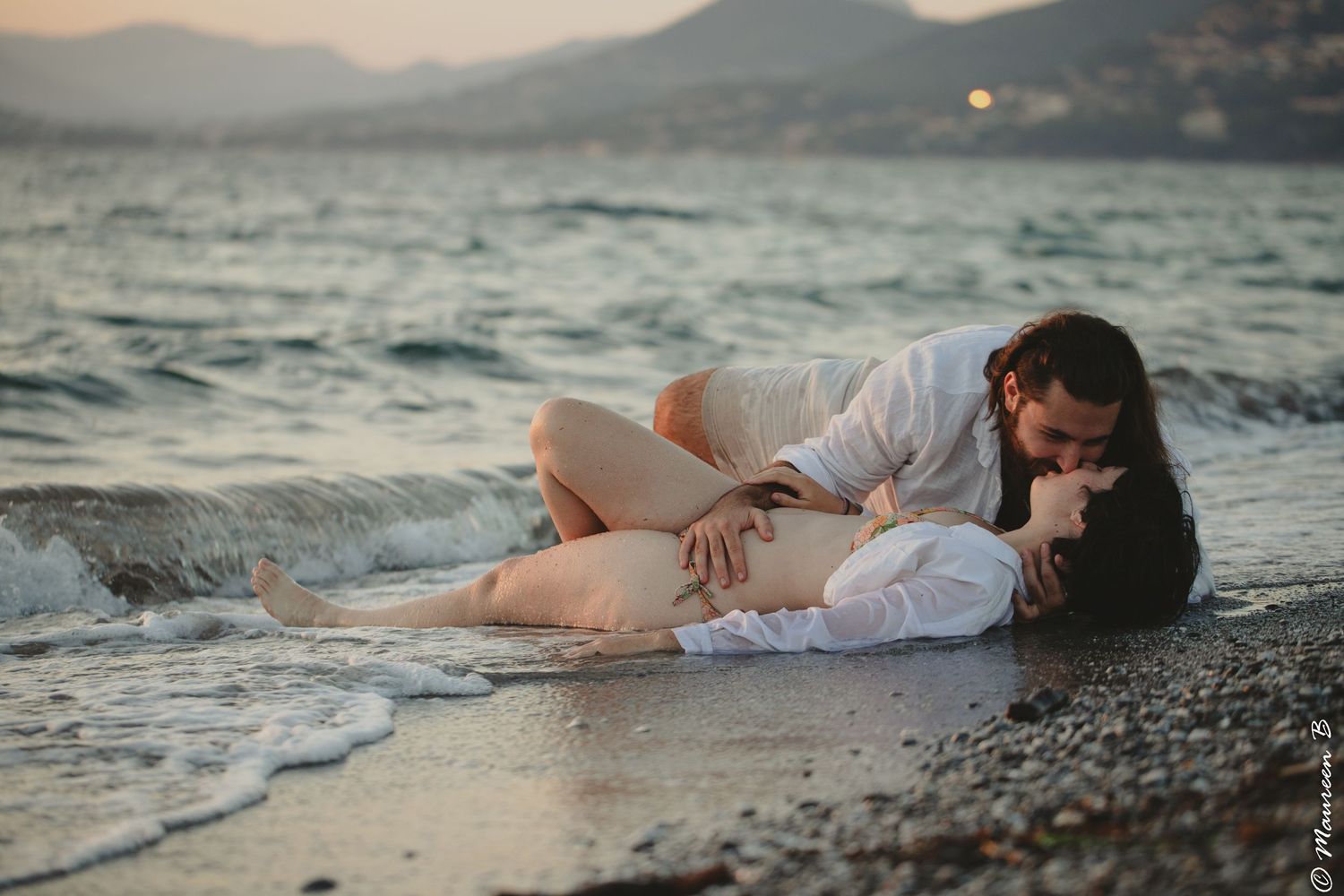 Romantic couple kissing while lying together on a beach shore at sunset.