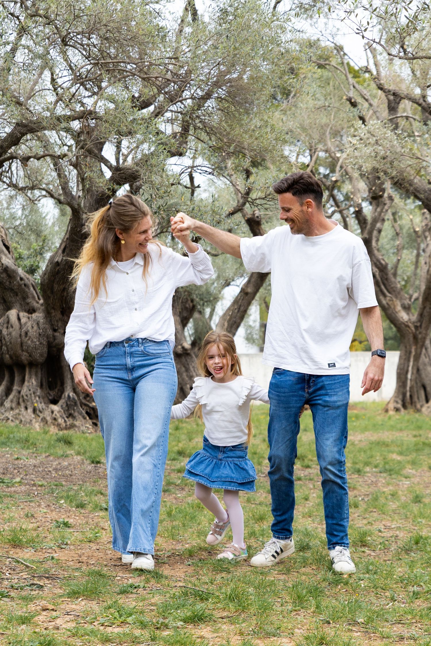 Happy family with young daughter swinging between parents in a scenic olive tree park.