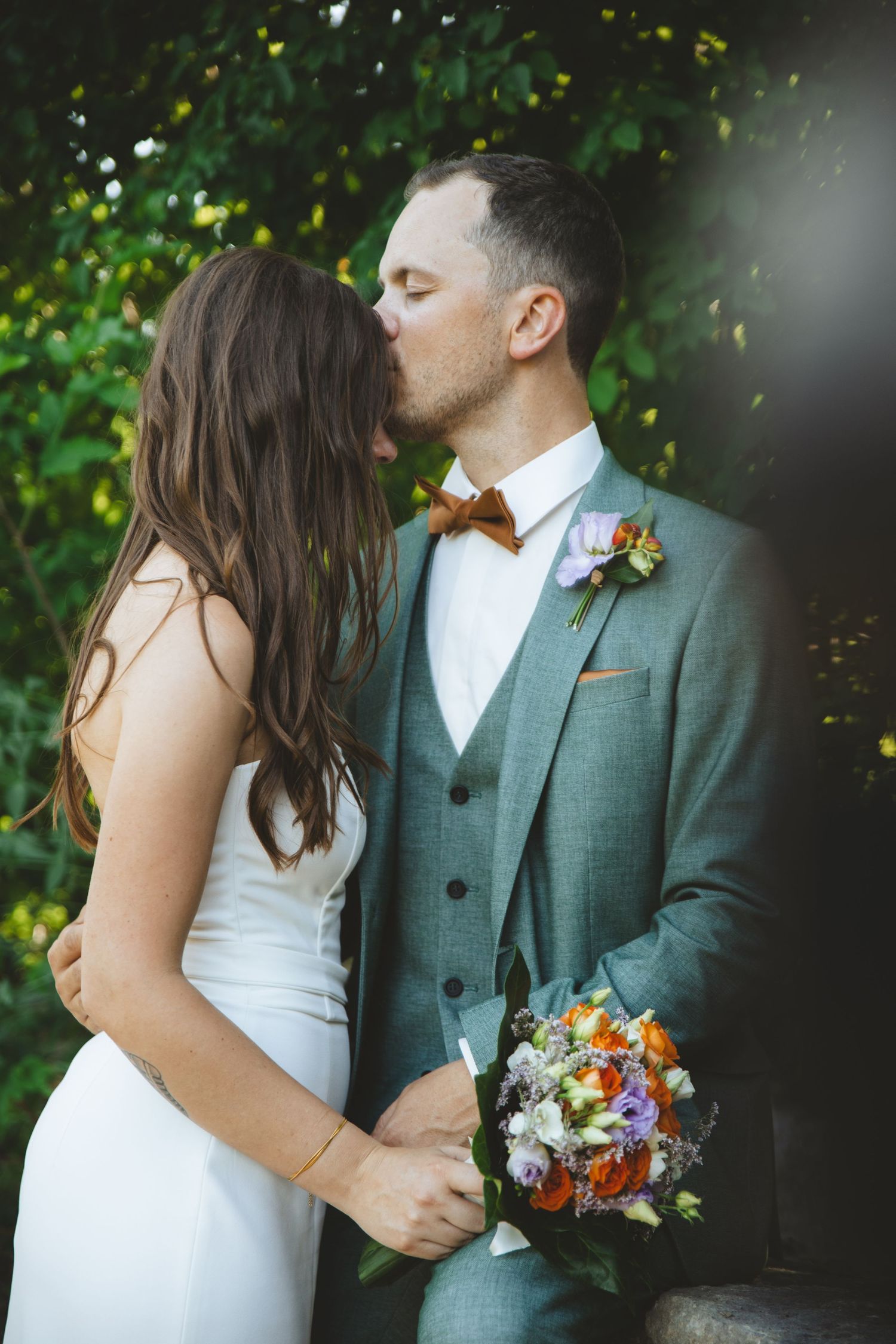 Groom kissing bride on forehead while holding bouquet against green leafy background outdoors.