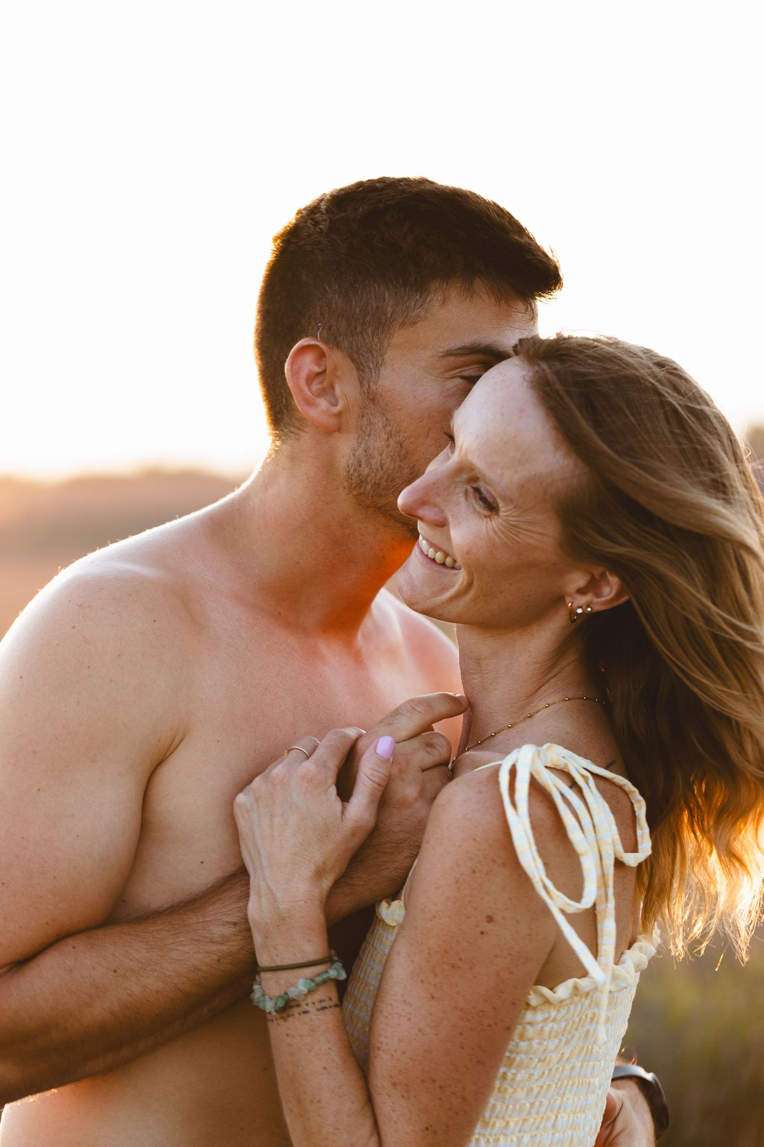 Loving couple embracing intimately at sunset, woman smiling joyfully as man kisses her cheek outdoors.
