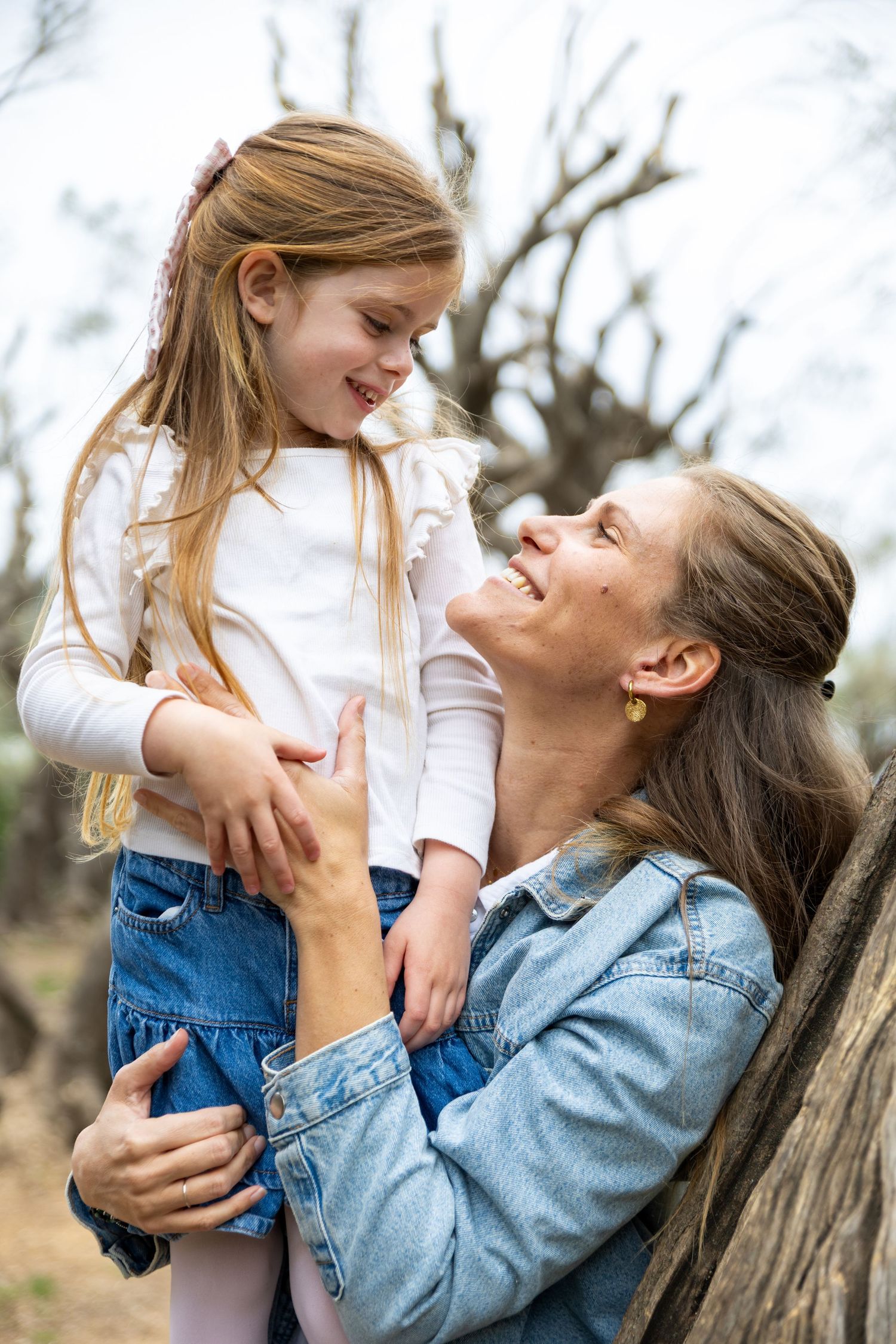 A smiling mother holds her young daughter close while sitting against a tree in a natural outdoor setting.