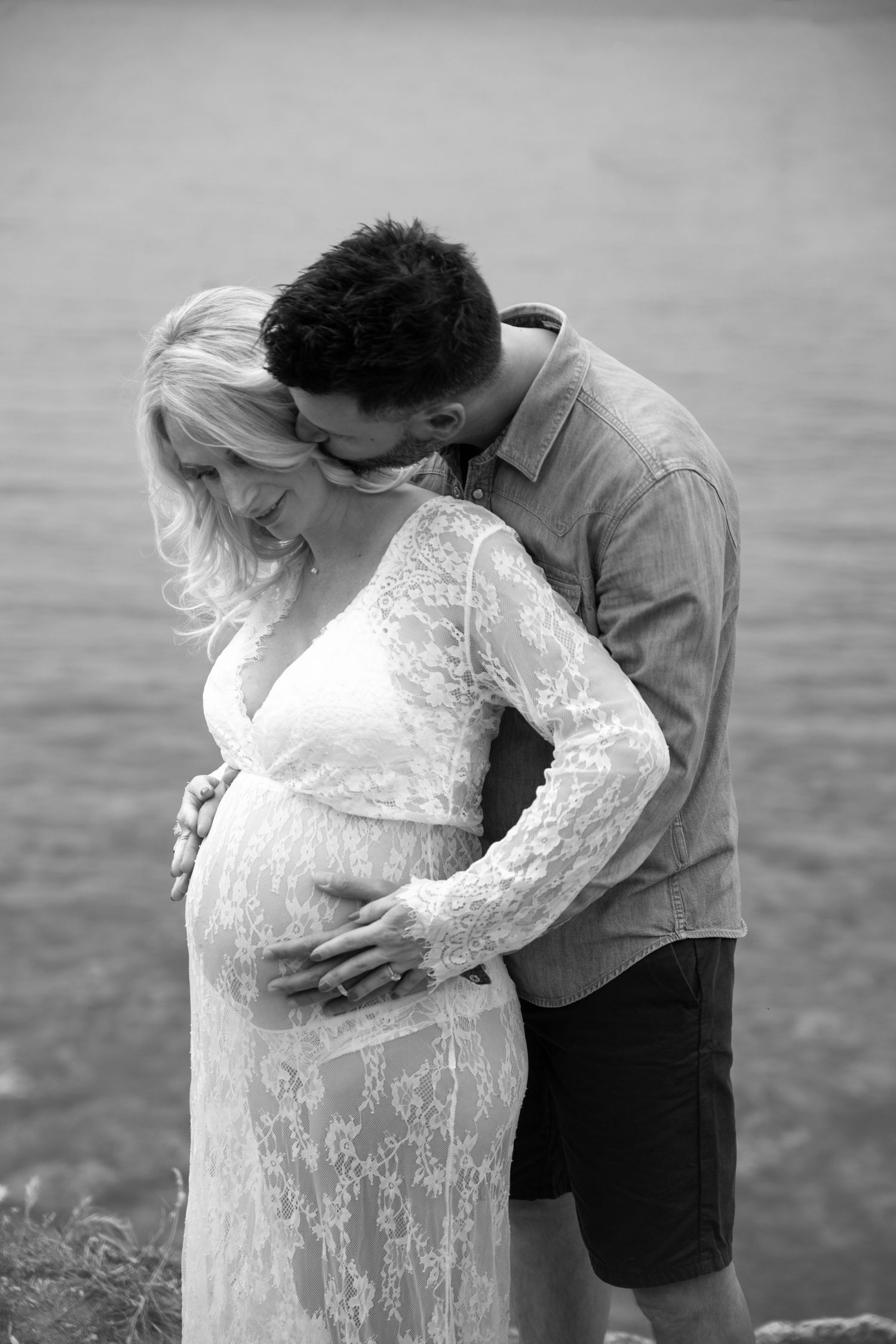 Black and white maternity photo of a couple embracing near water, man kissing woman's head while cradling her pregnant belly.