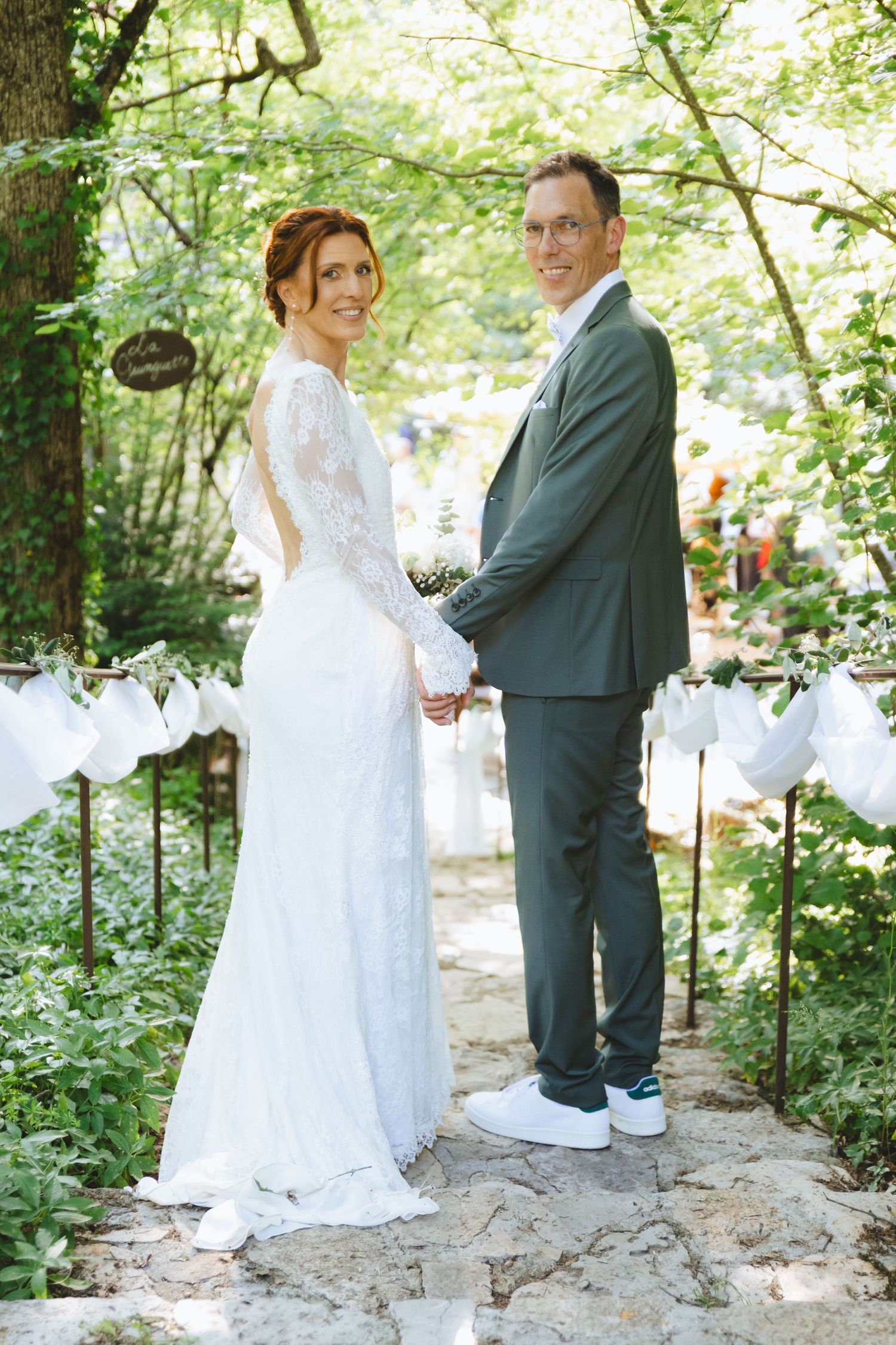 Bride in lace gown and groom in gray suit hold hands under green arch at outdoor garden wedding ceremony.