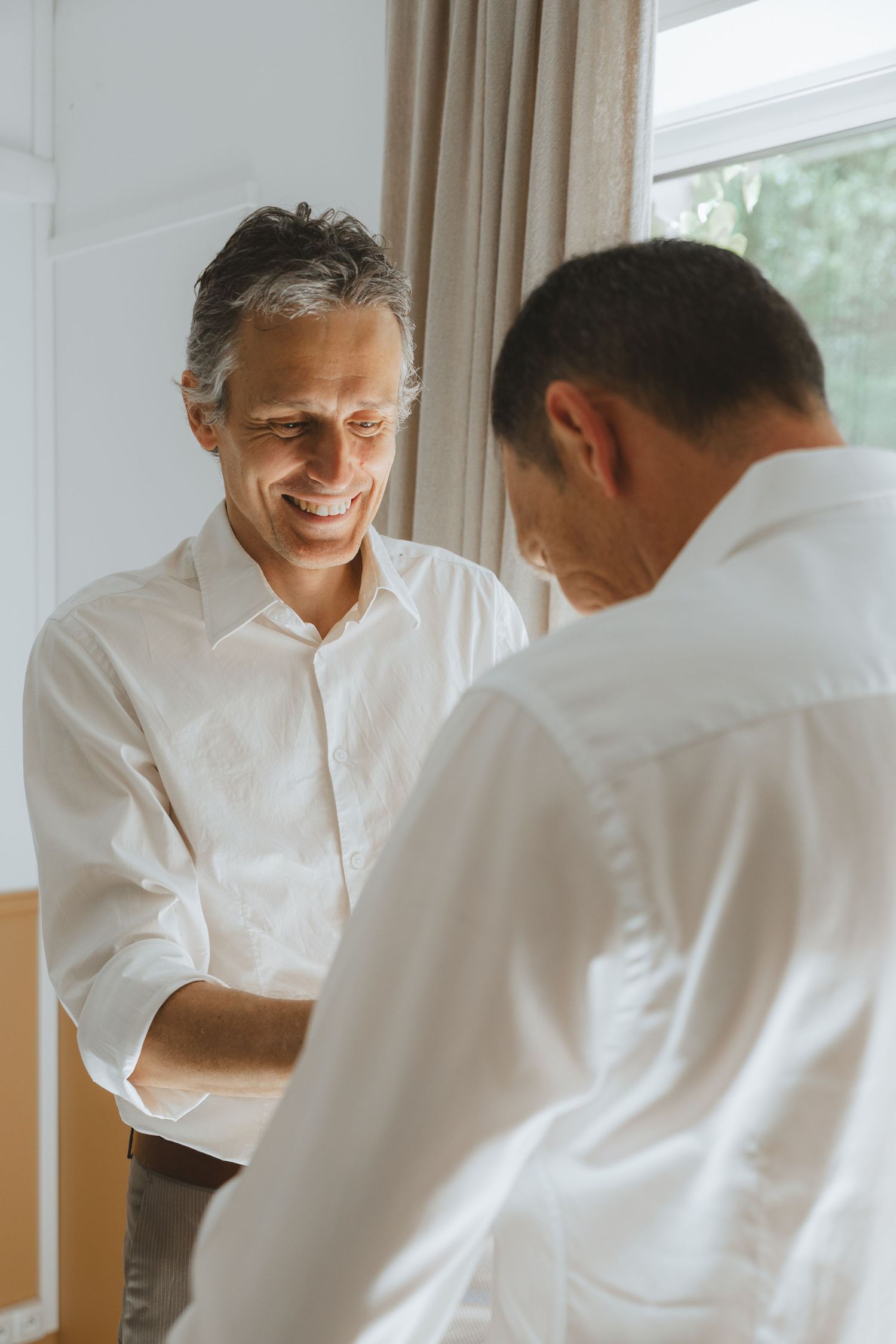 Two men in white shirts smiling and shaking hands in a bright room near a window.