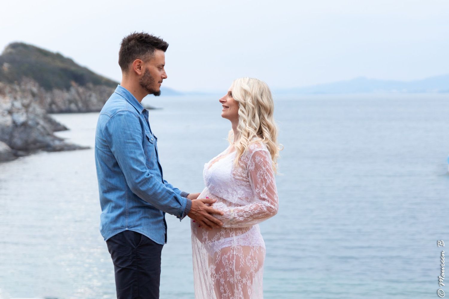 Couple standing by the sea, the pregnant woman in a sheer lace dress facing her partner.