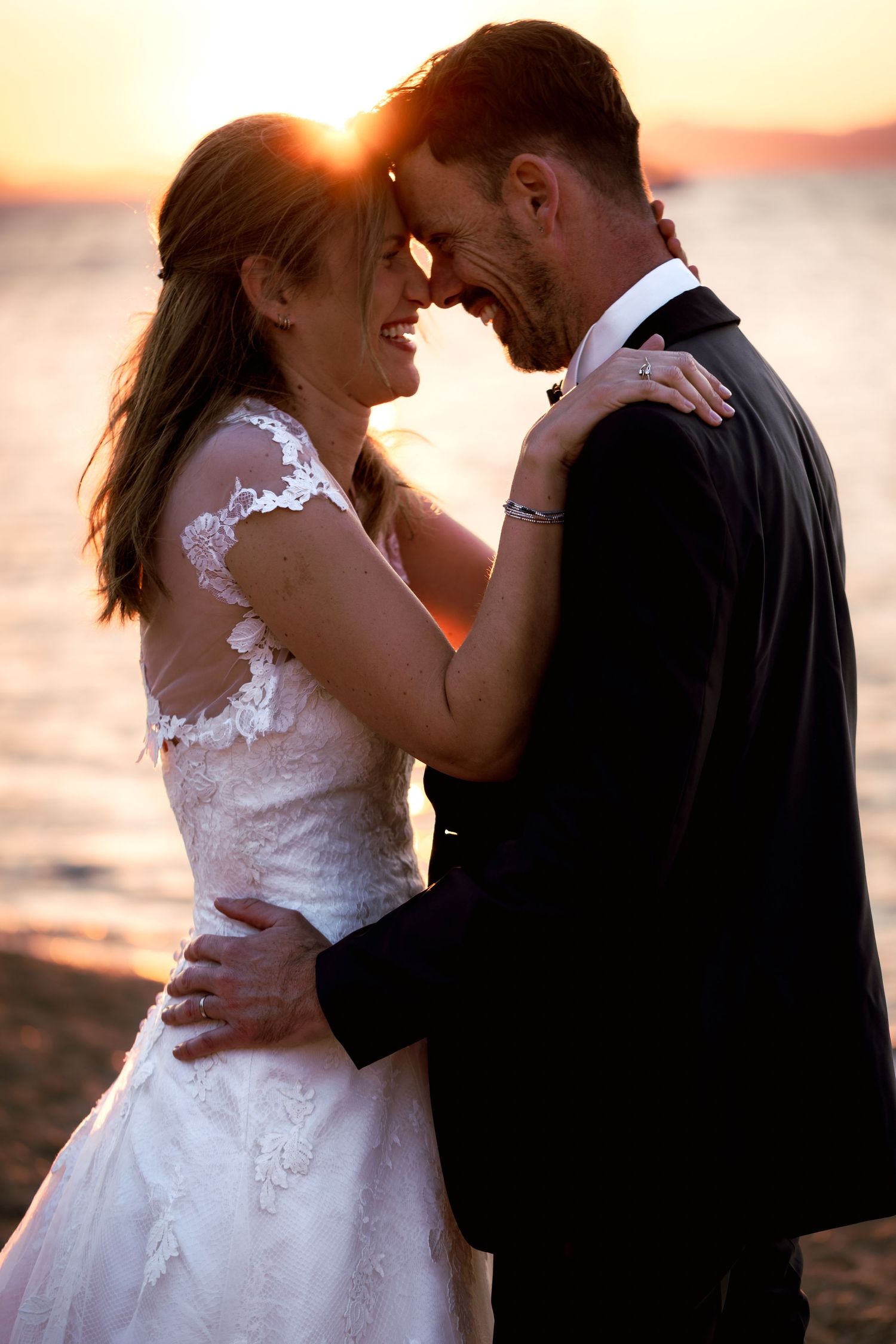 Bride and groom embracing on a beach at sunset, smiling with foreheads together in a romantic wedding moment.